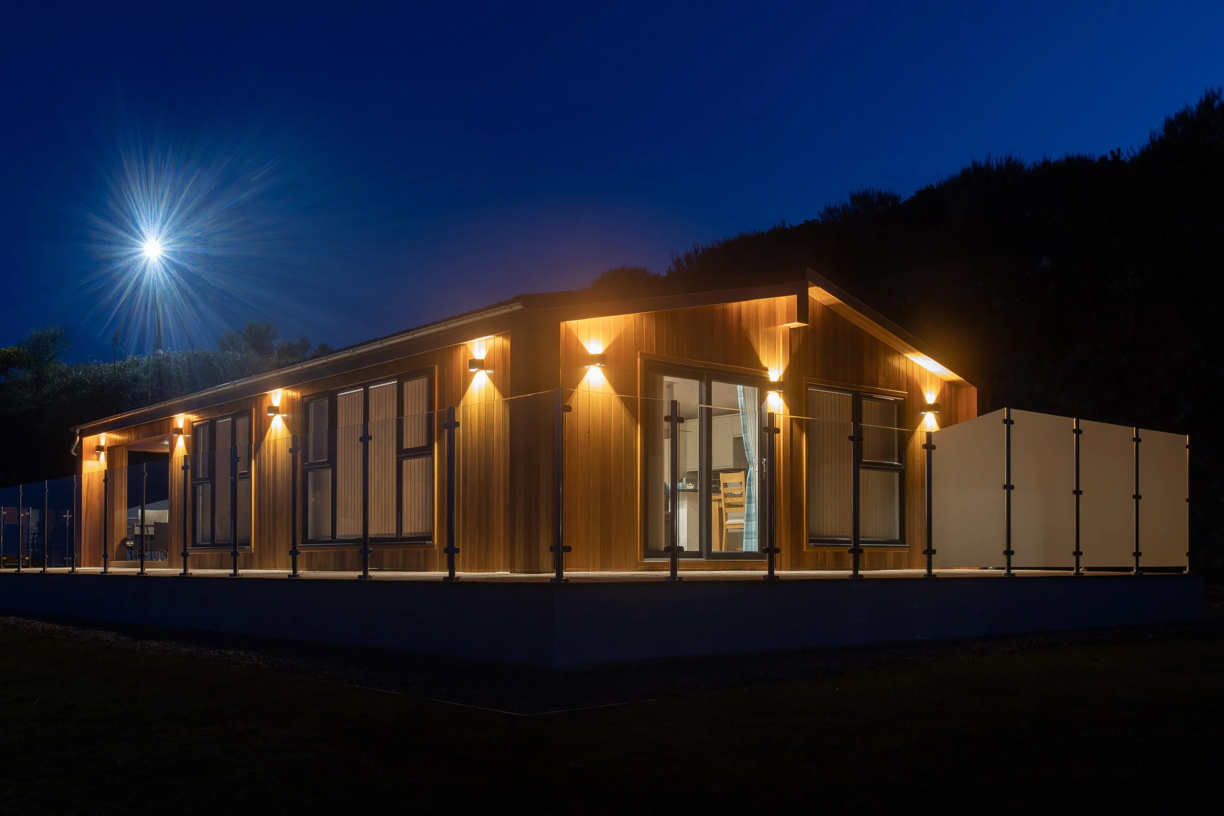A modern wooden house with large windows and outdoor lighting, situated on a raised platform at night under a clear starry sky.