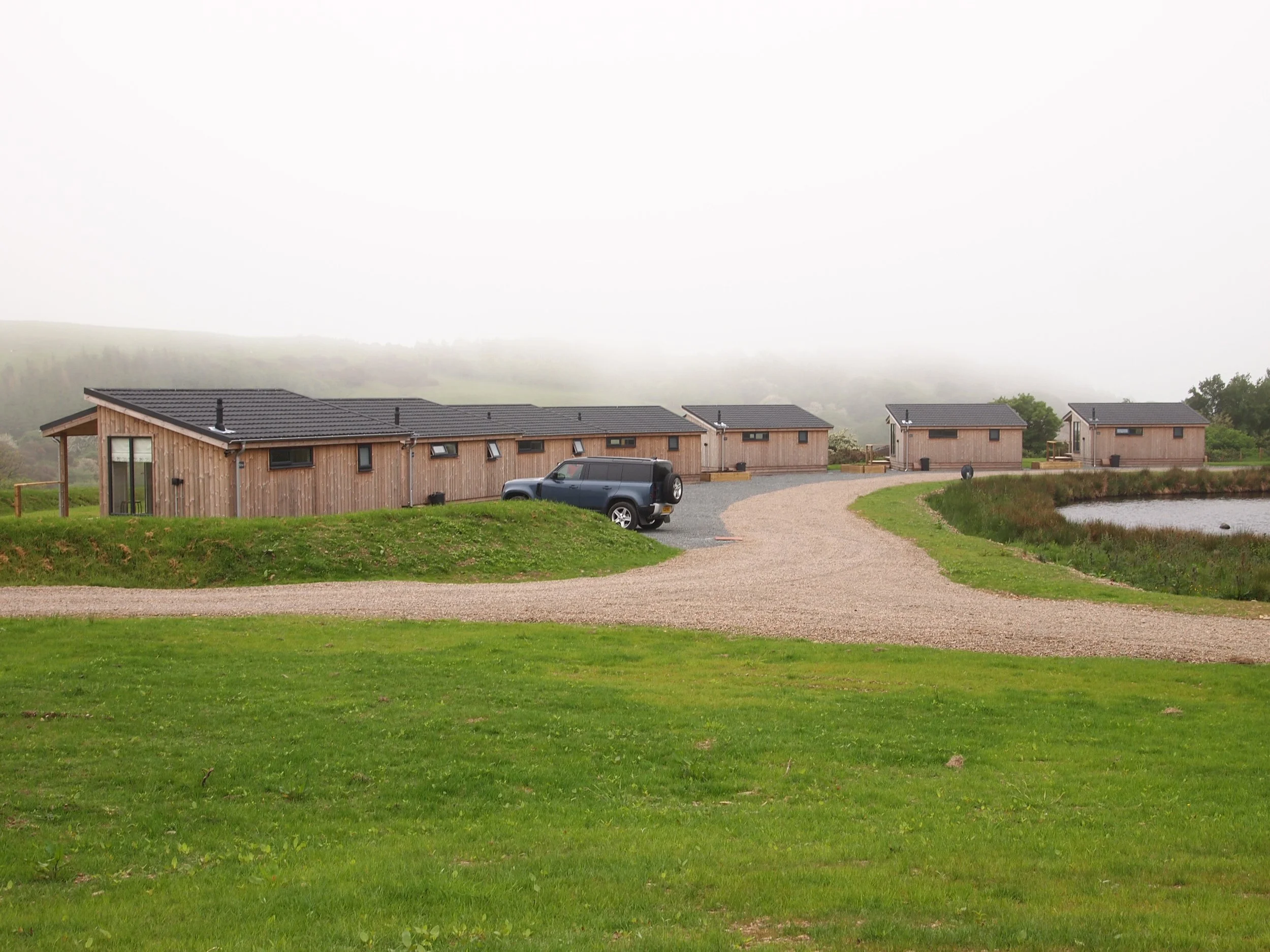 Several small wooden houses with black roofs near a pond, with a gravel pathway connecting them, on a foggy day.