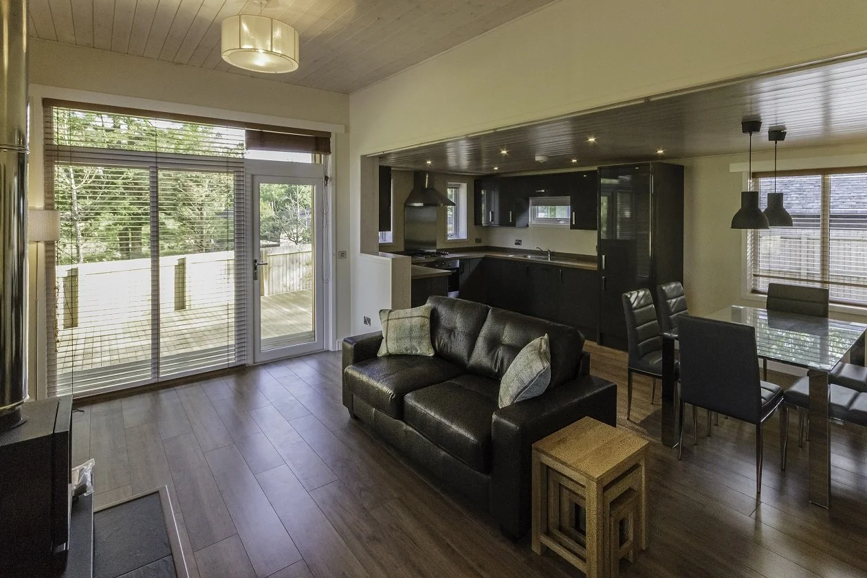 Living room with a black leather sofa, a wooden side table, a glass dining table with black chairs, a kitchen with black cabinets, and large windows with blinds overlooking an outdoor deck.