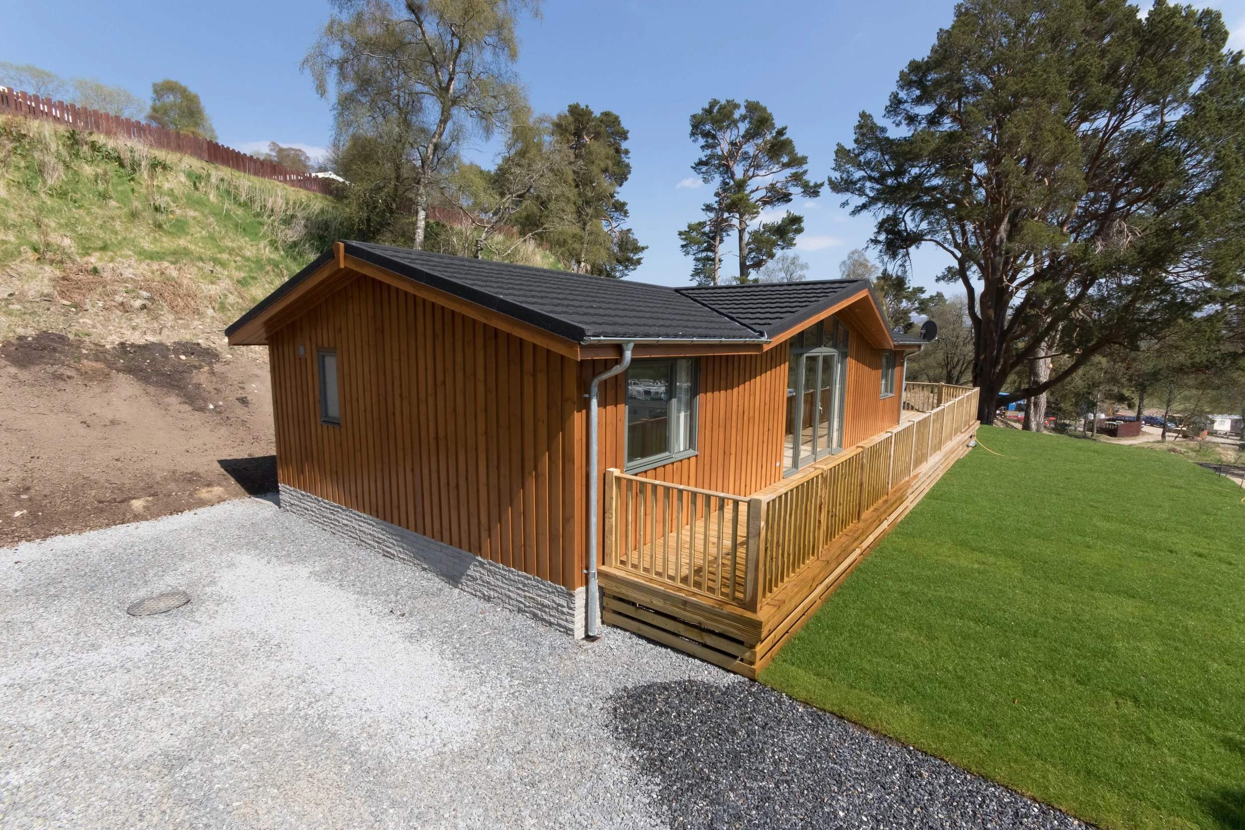 A wooden house with a black tiled roof and a small balcony, surrounded by grassy areas, trees, and a gravel driveway under a blue sky.