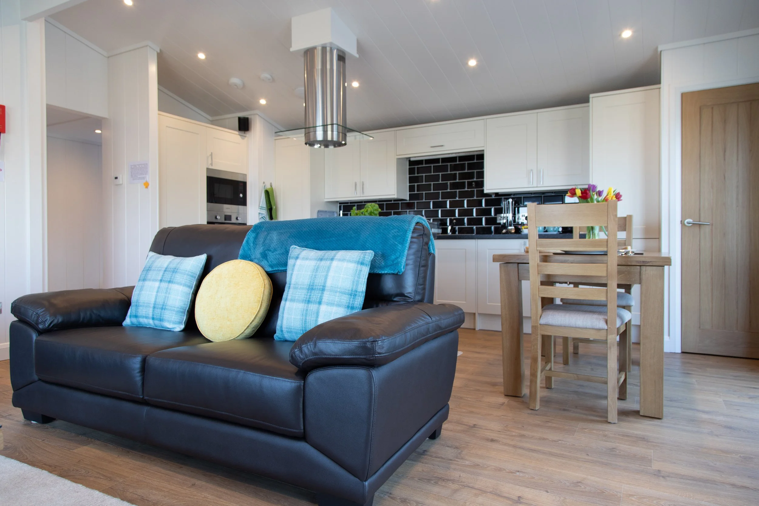 Living room with a black leather sofa, colorful cushions, and an open kitchen with white cabinets, black backsplash, and a wooden dining table with chairs.