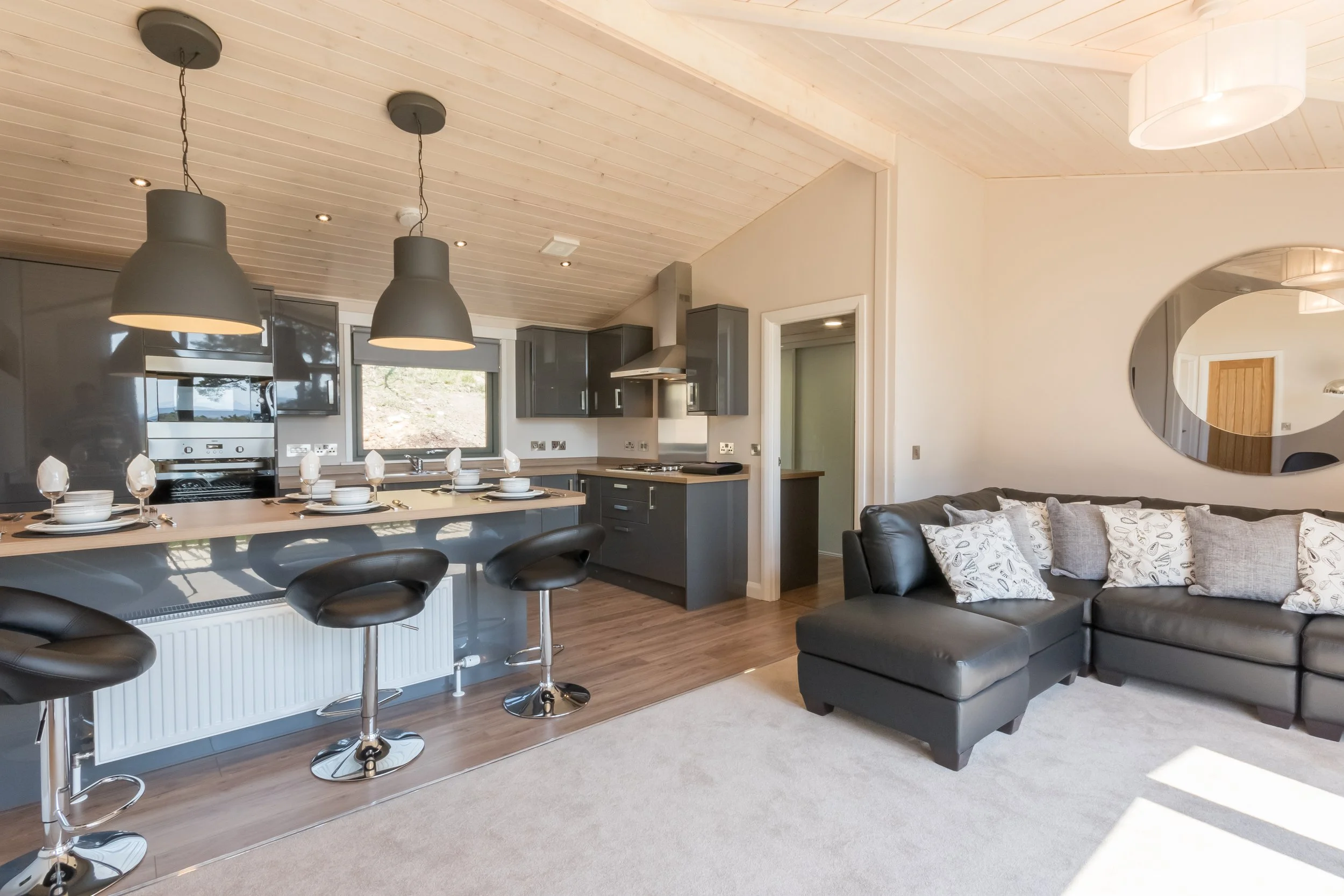 Open plan living room and kitchen with modern dark gray cabinetry, a kitchen island with bar stools, and a black leather sectional sofa with patterned throw pillows. There are two large pendant lights above the island and a window behind the sink.