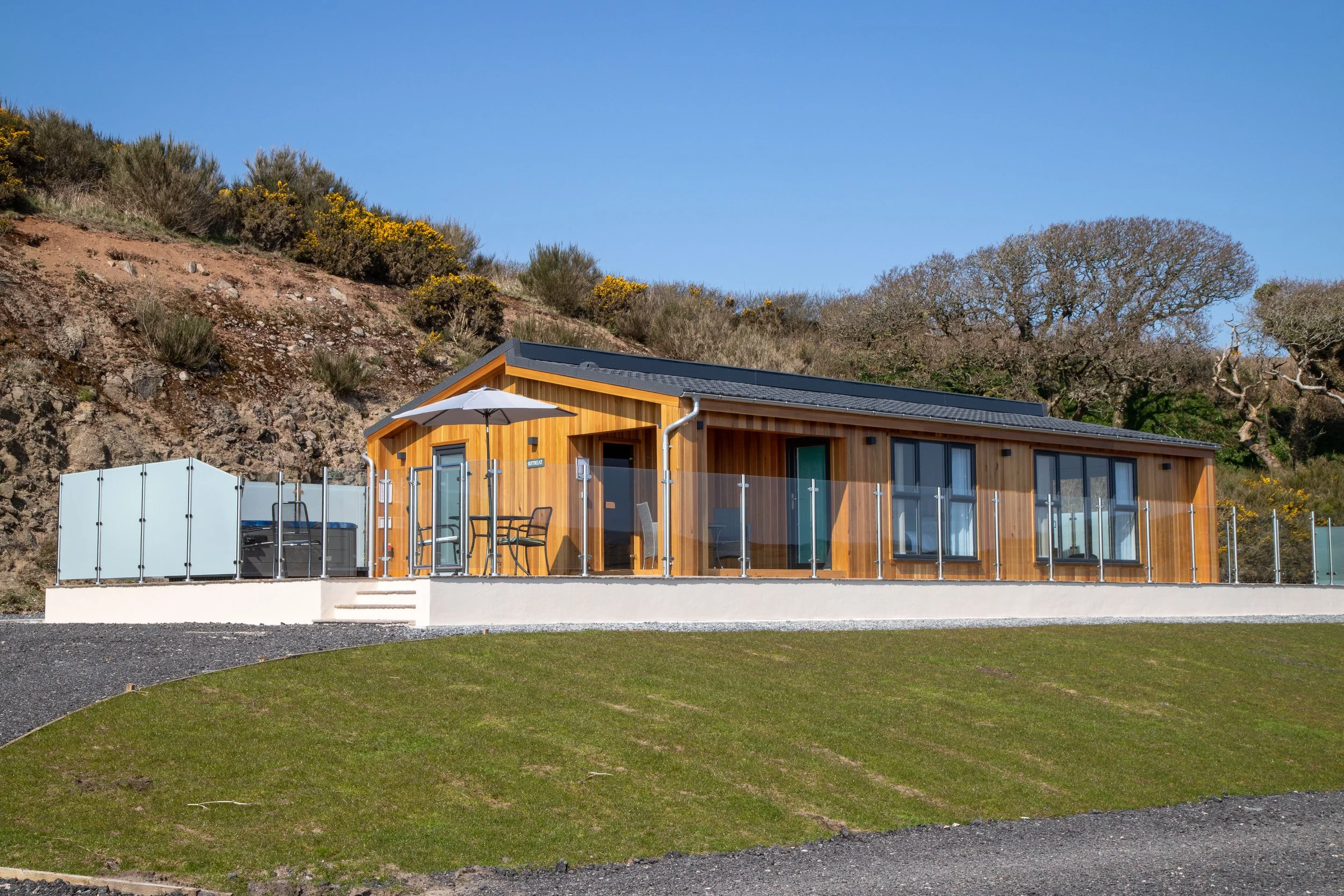 A modern wooden house with large glass windows and an outdoor patio with chairs and an umbrella, surrounded by a green lawn and hillside with trees and bushes under a clear blue sky.