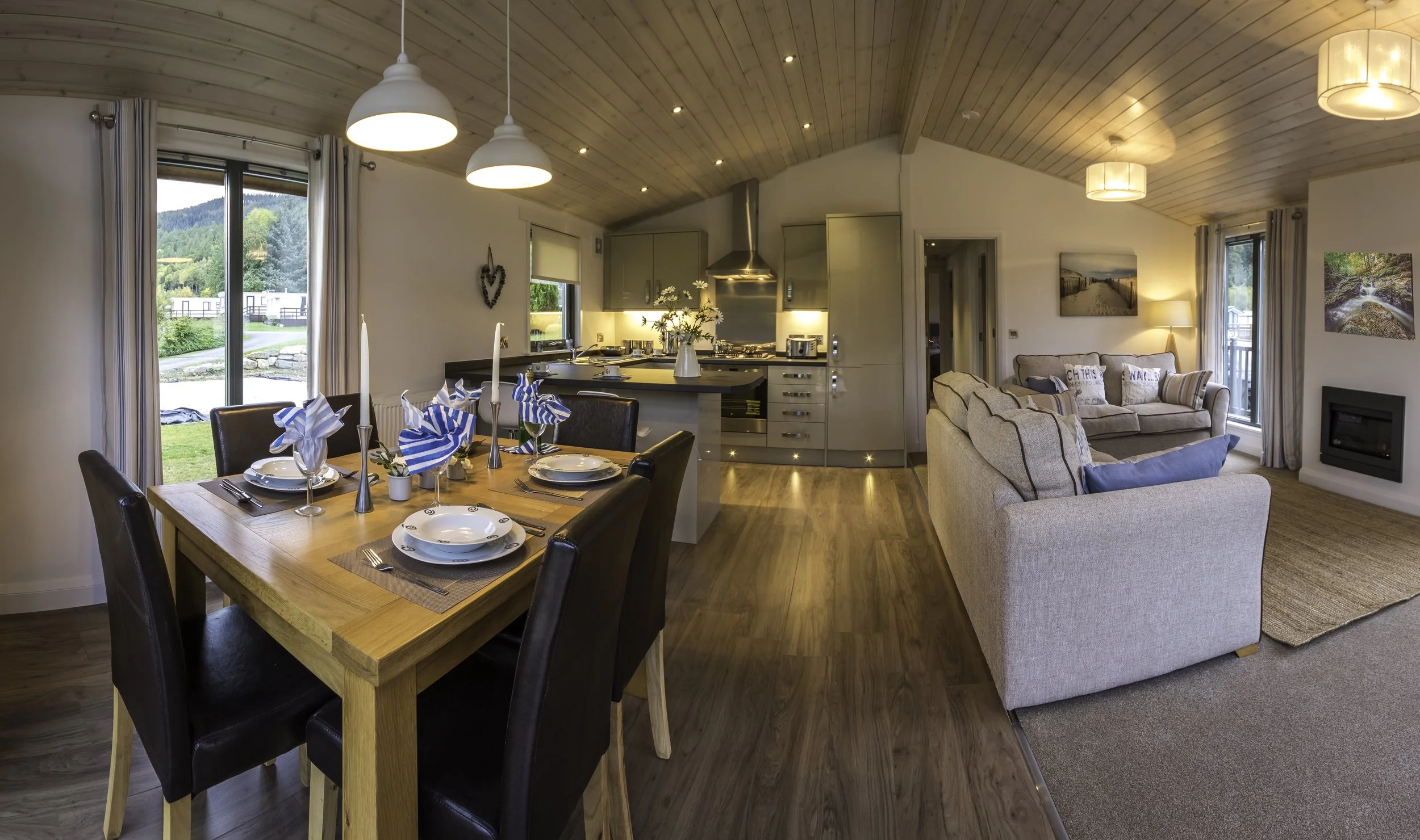 Open-concept living and dining area with a kitchen in the background, dining table set with plates, napkins, and candles, and a living room with couches, windows showing outdoor greenery.