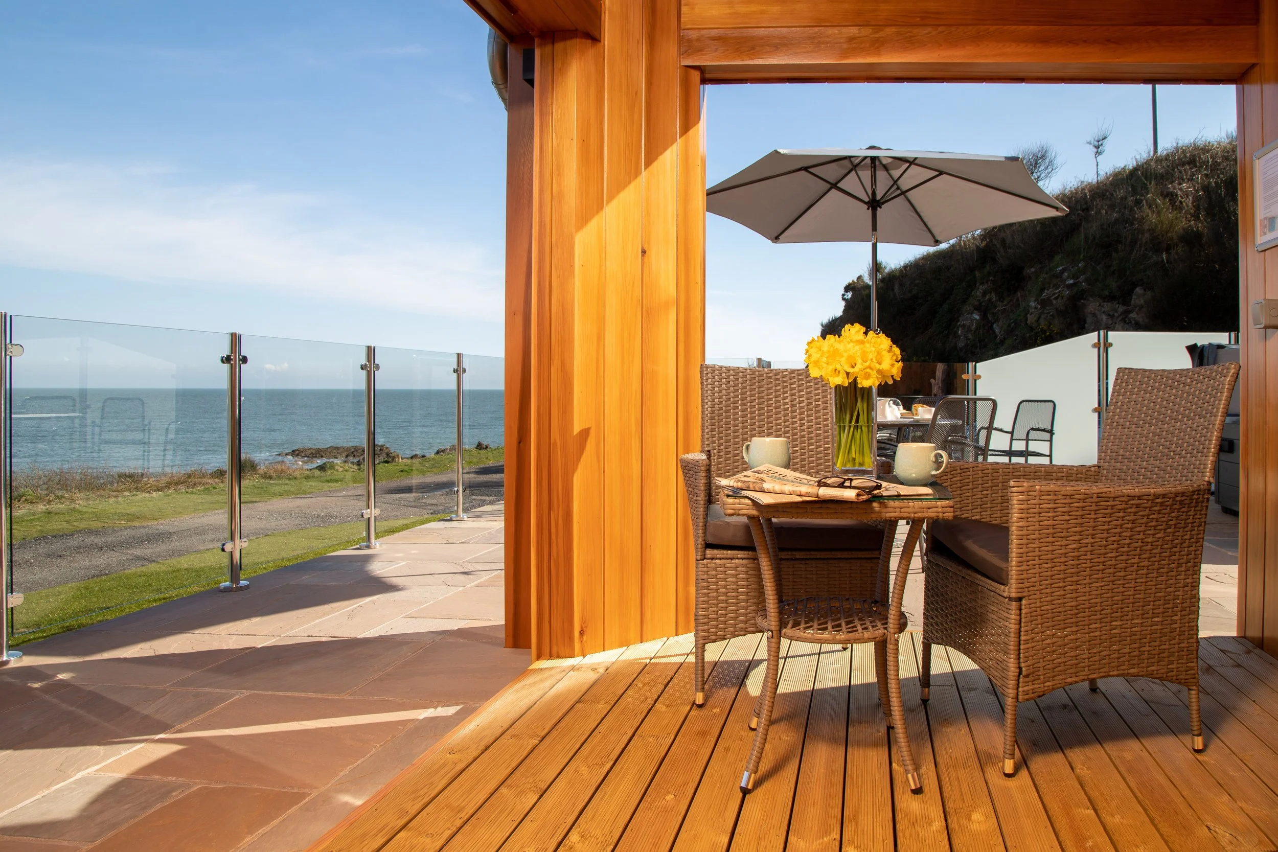 A cozy outdoor patio with wicker furniture, including a table with a vase of yellow flowers, two cups, and a newspaper, overlooking the ocean under a clear blue sky.