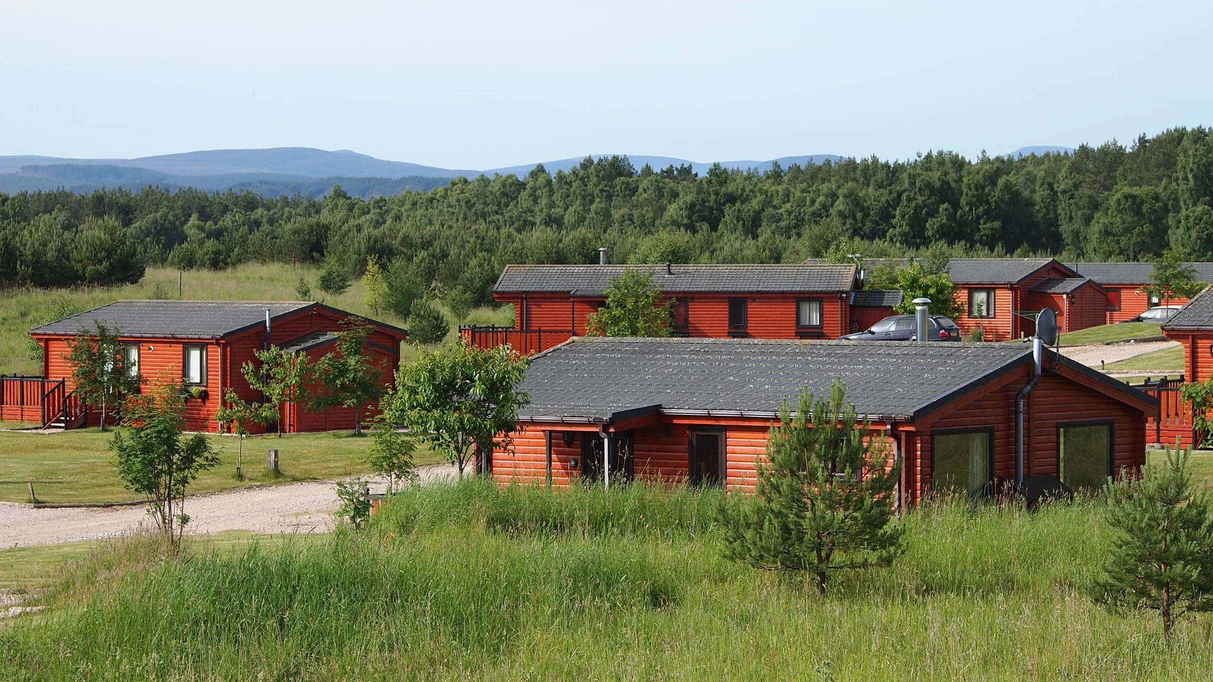 Multiple red wooden cabins with dark grey roofs in a grassy landscape with trees and distant mountains in the background.
