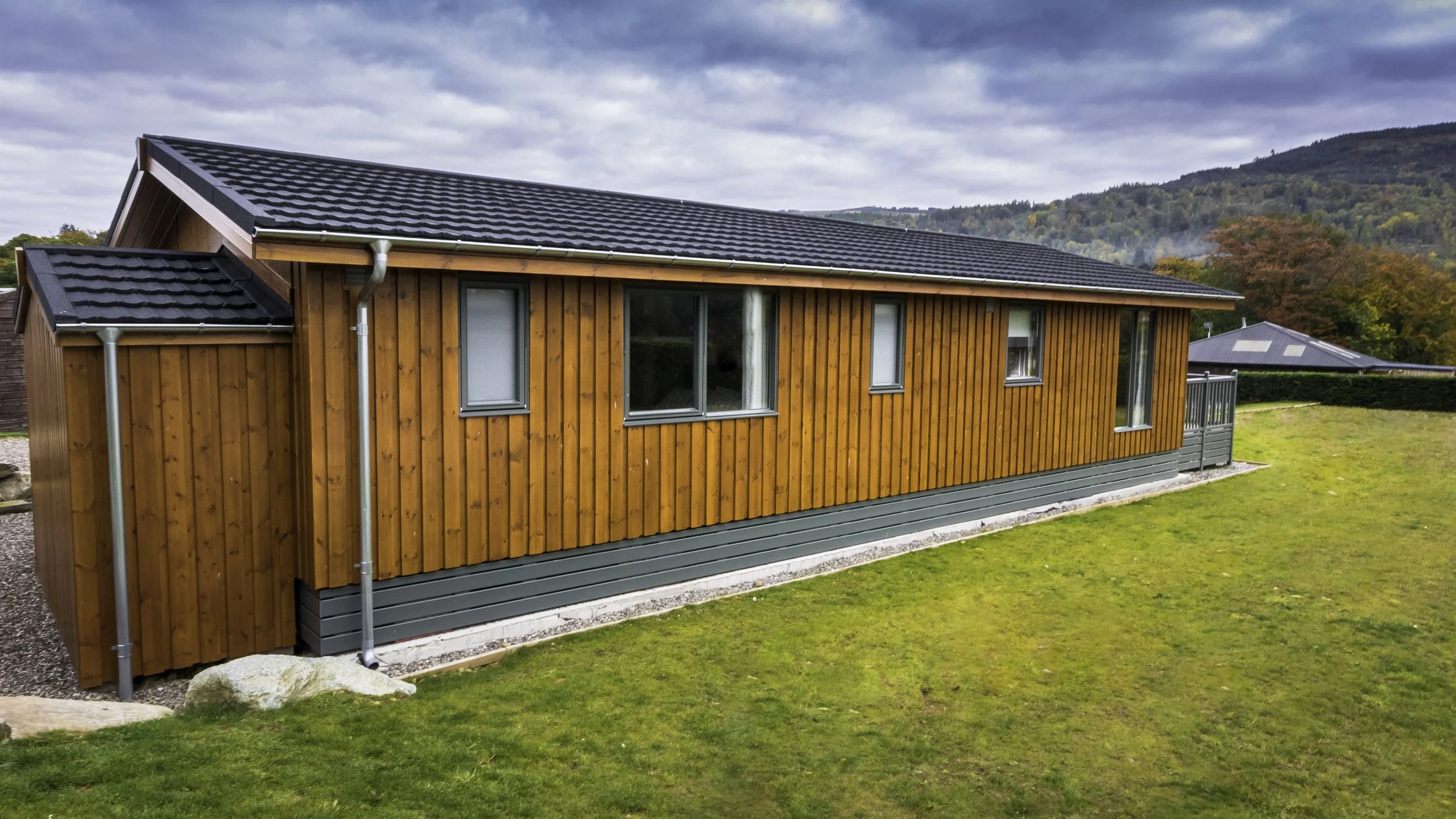 A wooden house with a black tile roof and multiple windows, situated in a grassy yard with a backdrop of trees and hills under a cloudy sky.
