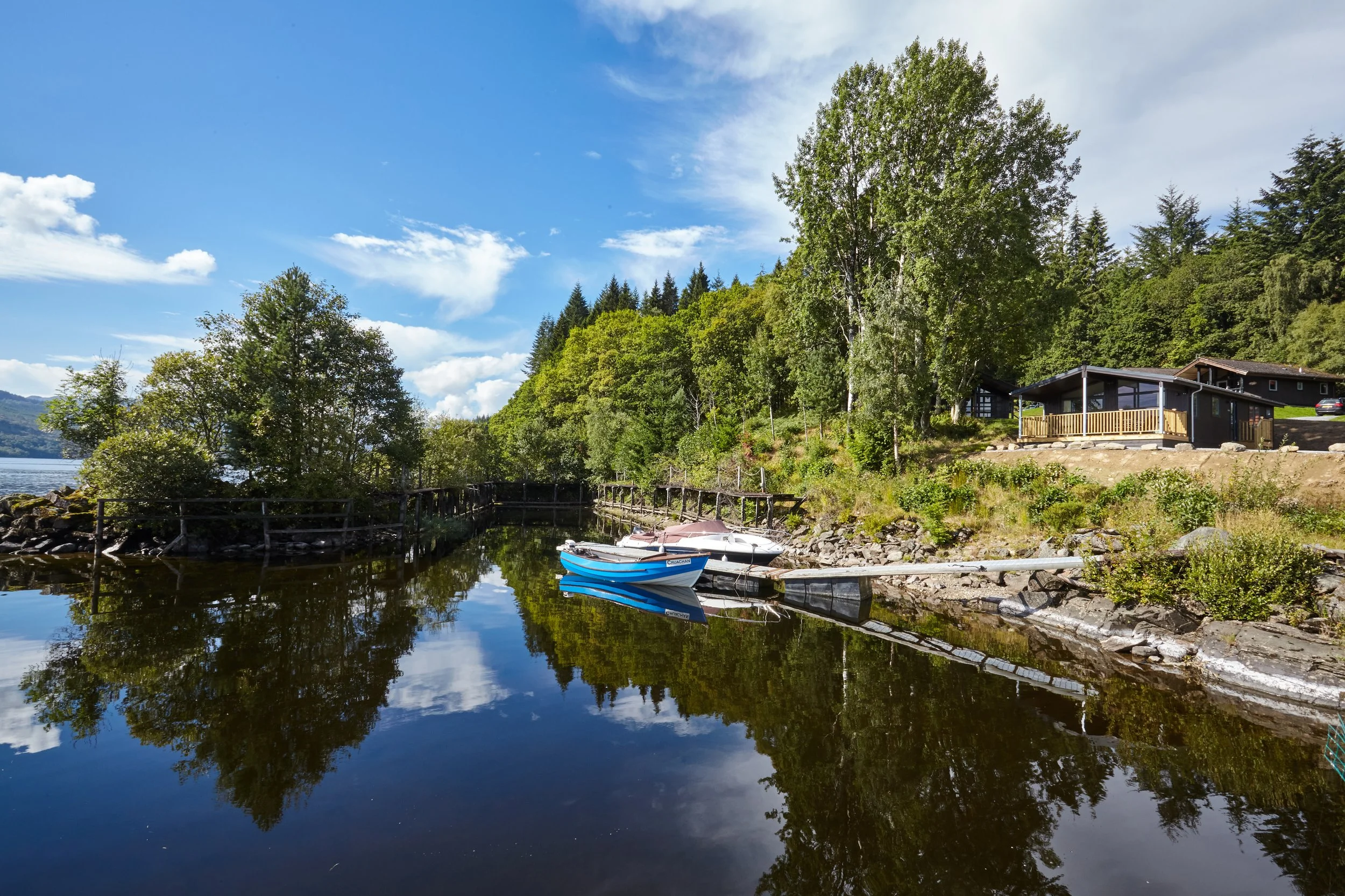 A peaceful lakeside scene with calm water reflecting trees and a house, with boats docked along the shore on a sunny day.