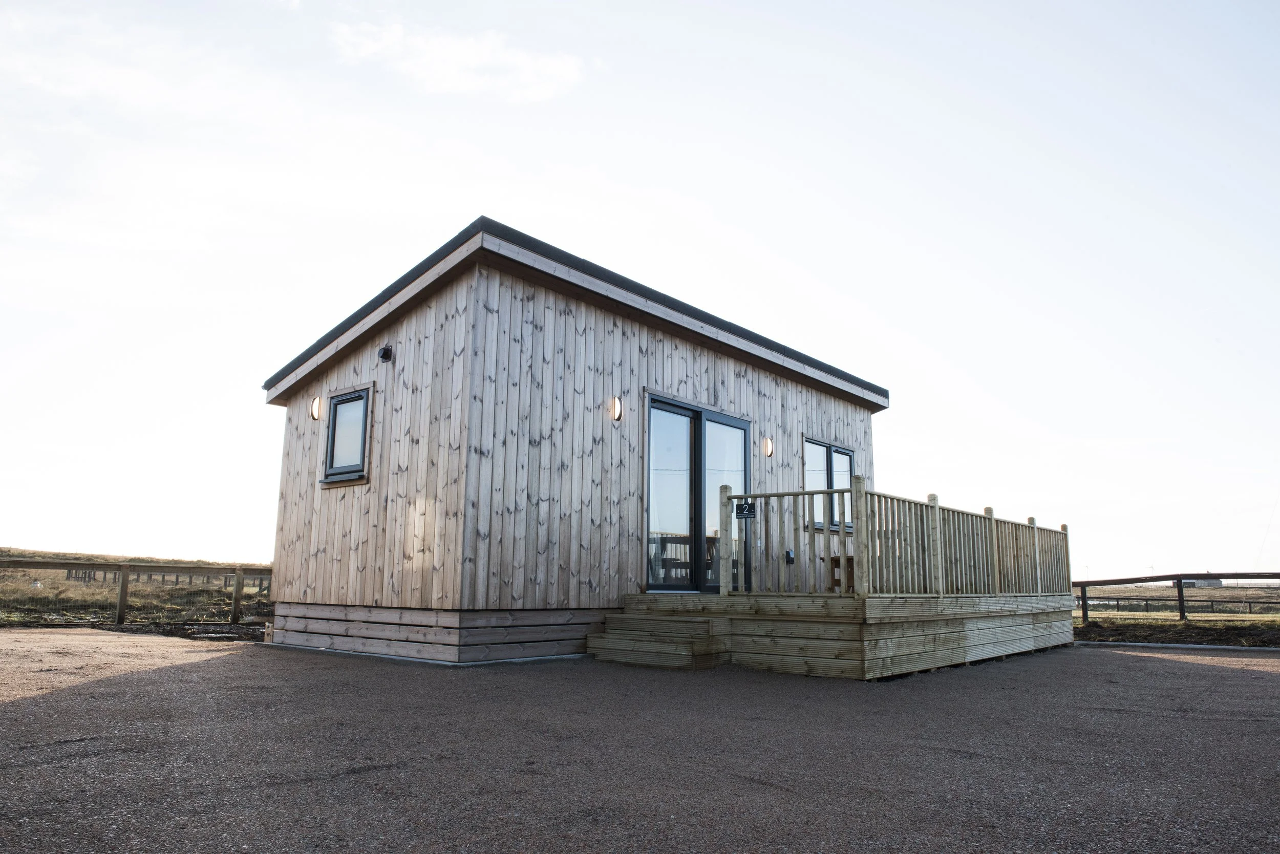 Small wooden house with a deck, located in a rural or open area under a clear sky.