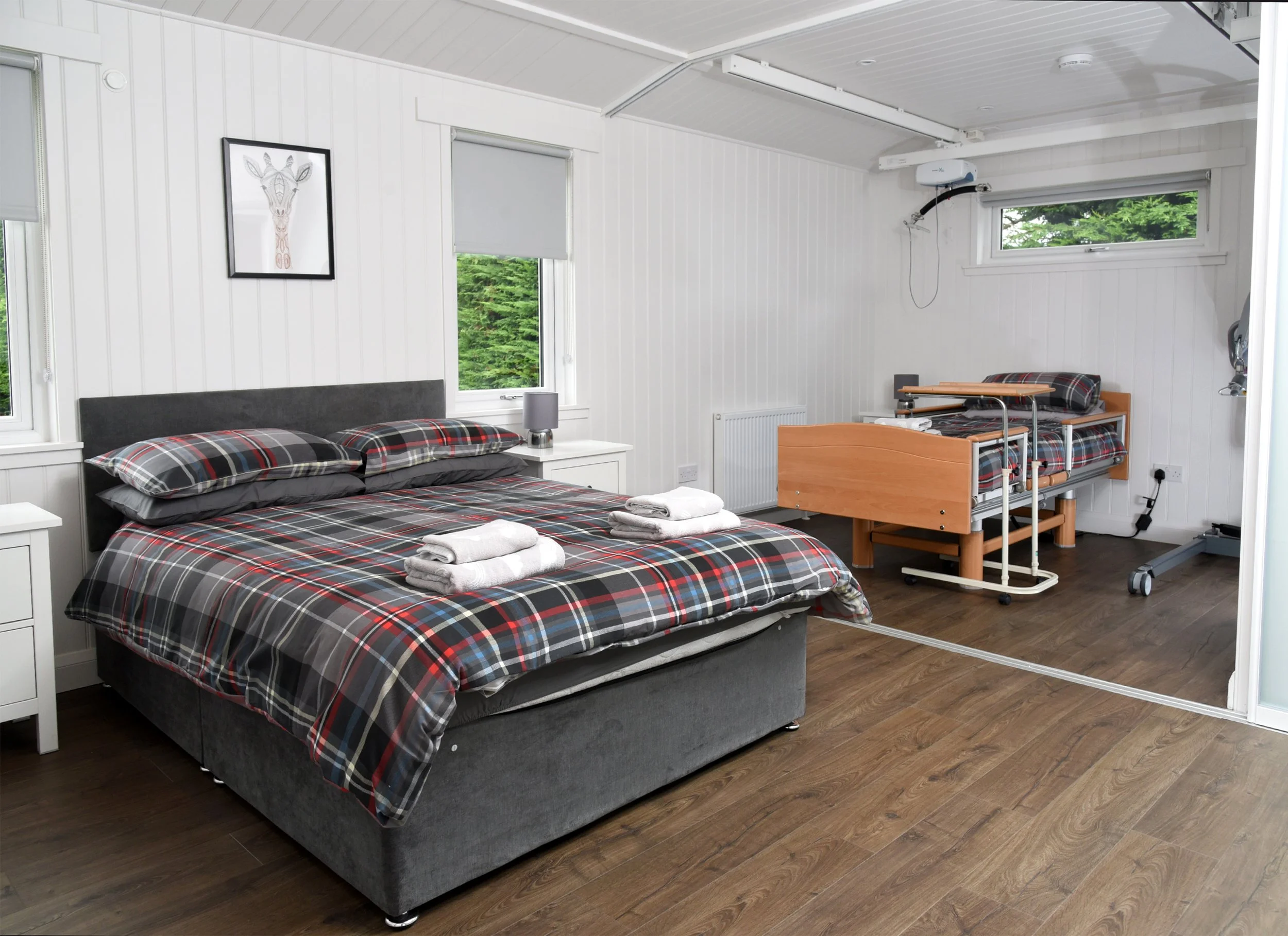 A hospital room with a large bed covered with a plaid duvet and pillows, a bedside table with a lamp, and a second bed designed for a patient with mobility aids, positioned near windows with greenery outside.