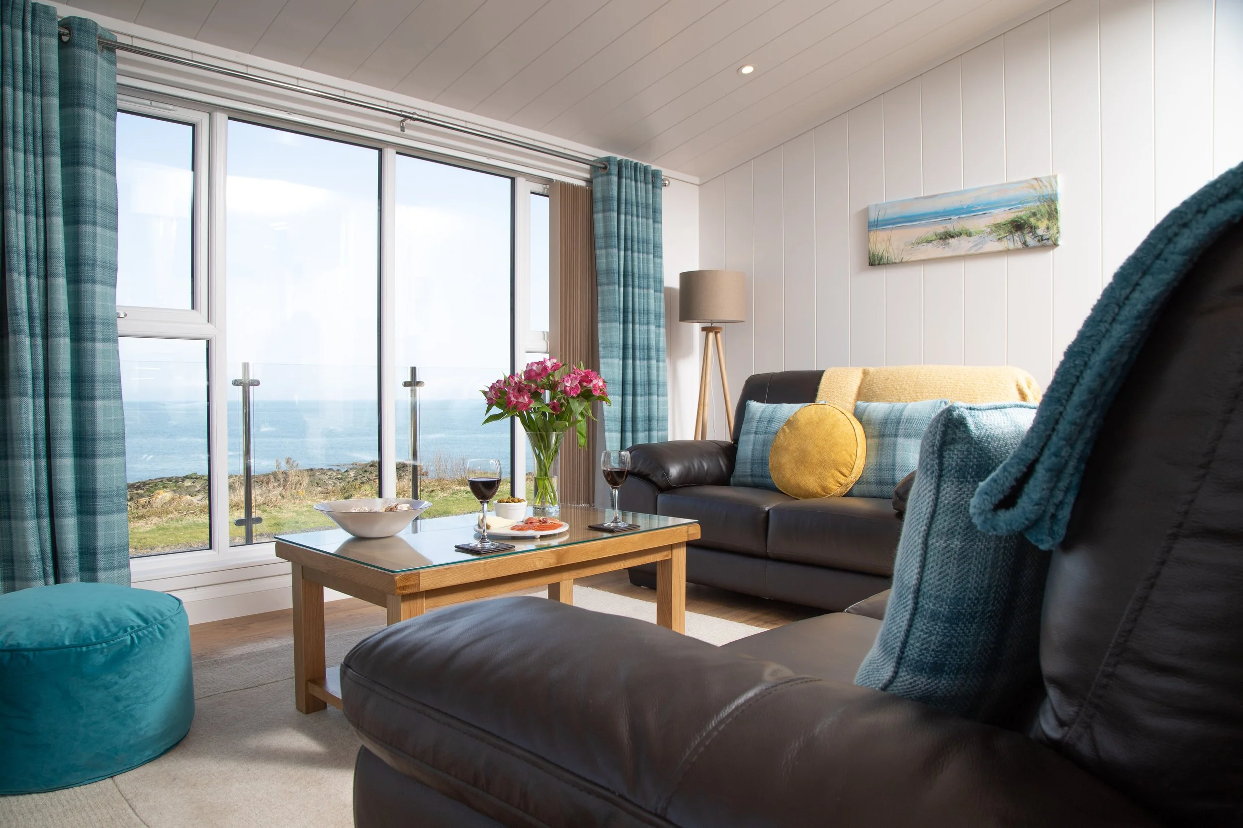 Living room with large windows overlooking the ocean, decorated with blue and yellow accents, a wooden coffee table with wine glasses and snacks, and a sofa with a yellow pillow and patterned cushions.