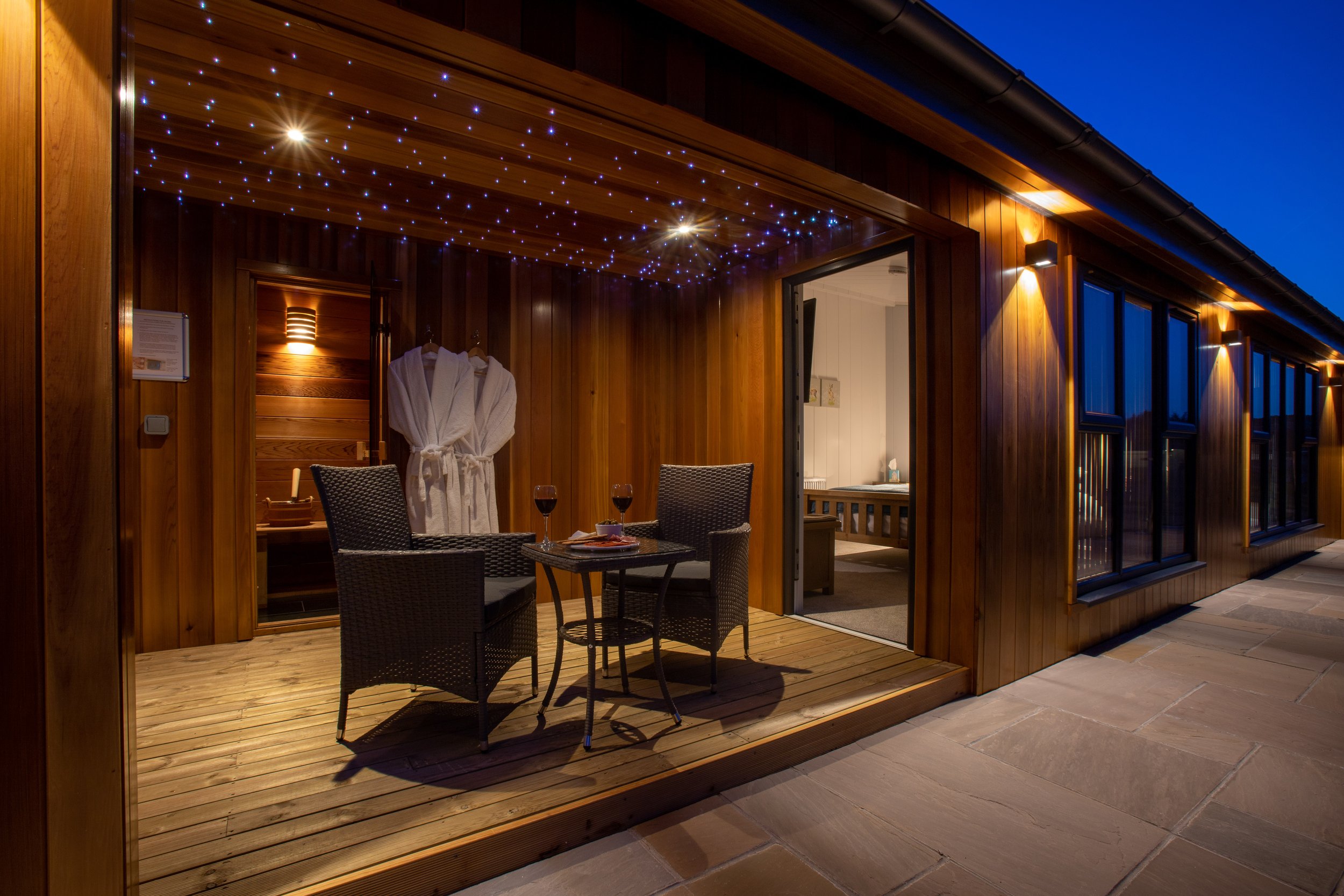 A cozy wooden patio at night featuring a small table with two wine glasses, two chairs, and a robe hanging inside a sauna or shower area illuminated by string lights.