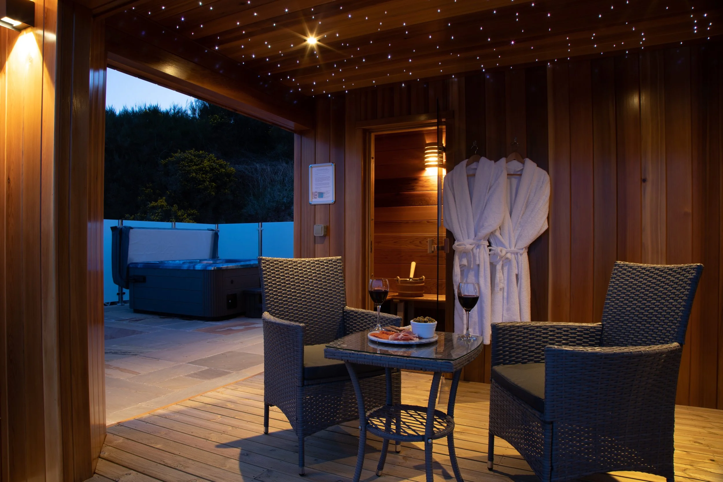 An outdoor patio area with wooden walls and ceiling, decorated with star-shaped lights. There are two bathrobes hanging on a wall, a small table with two glasses of red wine, a bowl of snacks, and a plate with food. The patio opens to a hot tub outsi