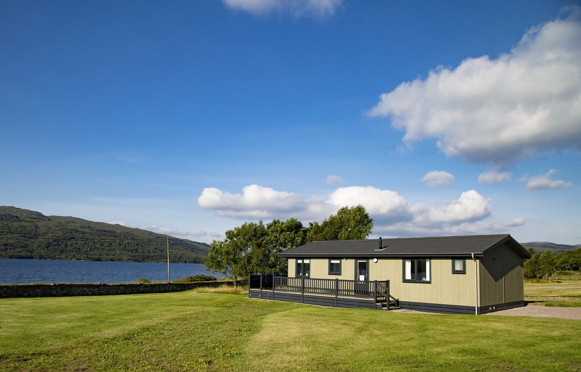 A house with a black deck and railing, situated on a grassy field near a body of water with hills in the background, under a partly cloudy blue sky.