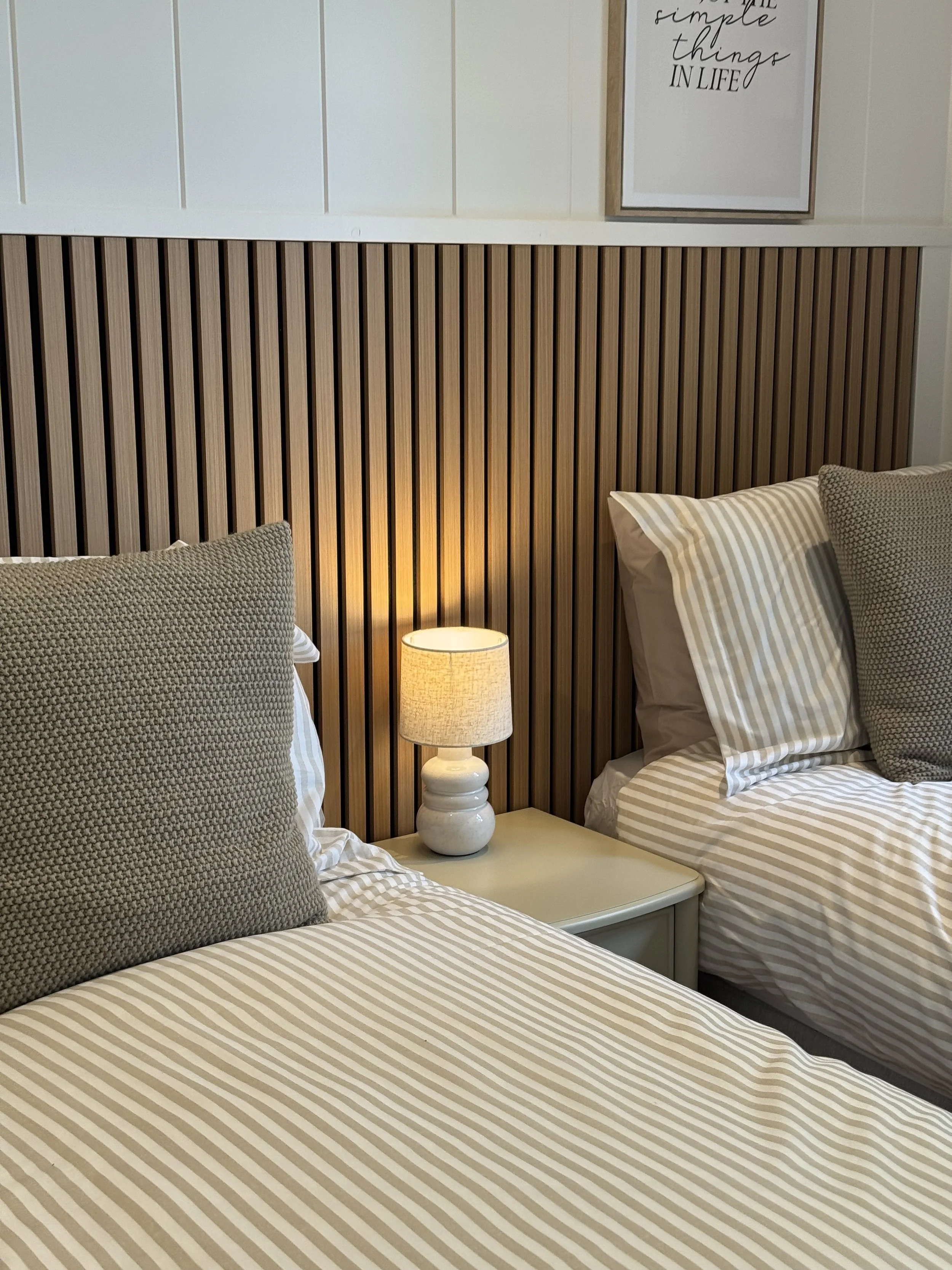 Two beds with beige and white striped bedding and textured beige pillows, a white nightstand with a lit table lamp, and a wooden slat headboard against a white paneled wall with framed wall art.