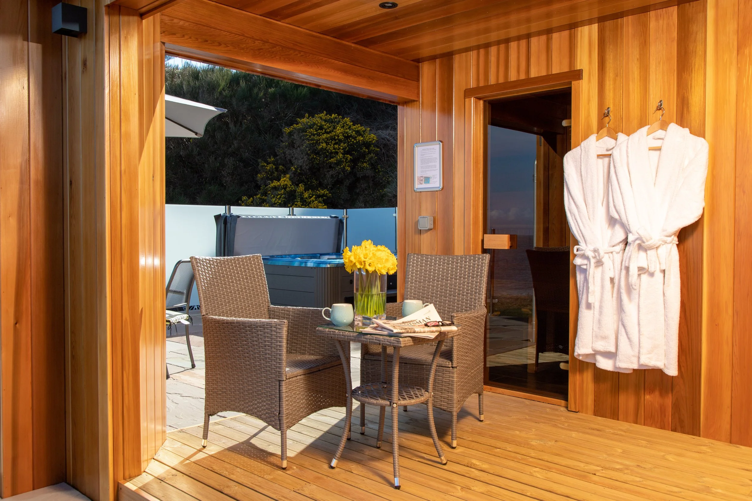 Outdoor wooden deck with chairs, table, journal, flowers, and robes, overlooking a landscape with trees and sky.