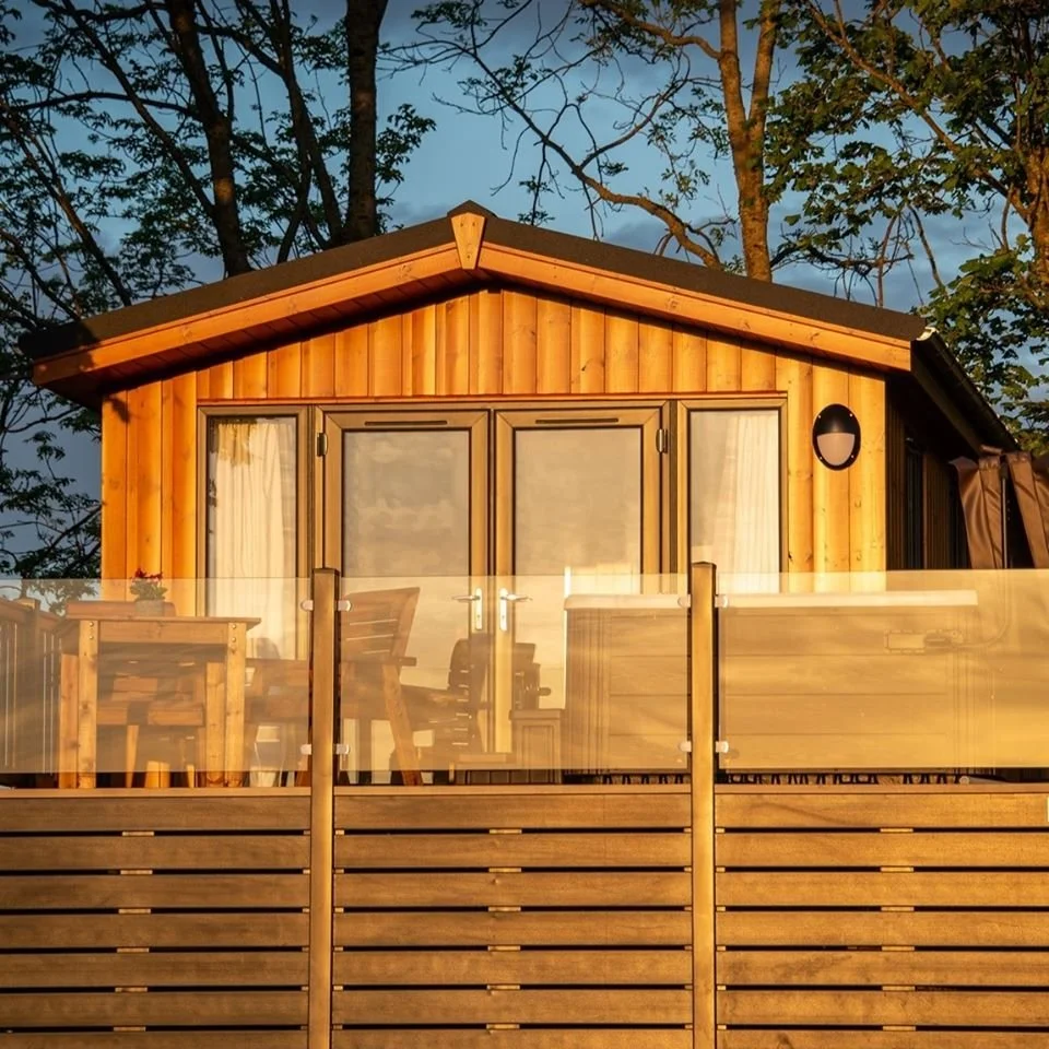 A wooden cabin with large sliding glass doors and a balcony with outdoor furniture, surrounded by trees at sunset.