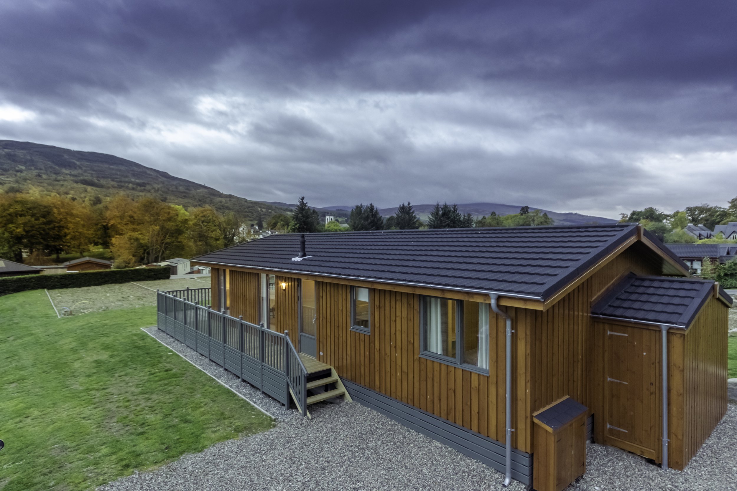 A modern wooden house with a black tiled roof, situated in a scenic landscape with hills and trees, during overcast weather.