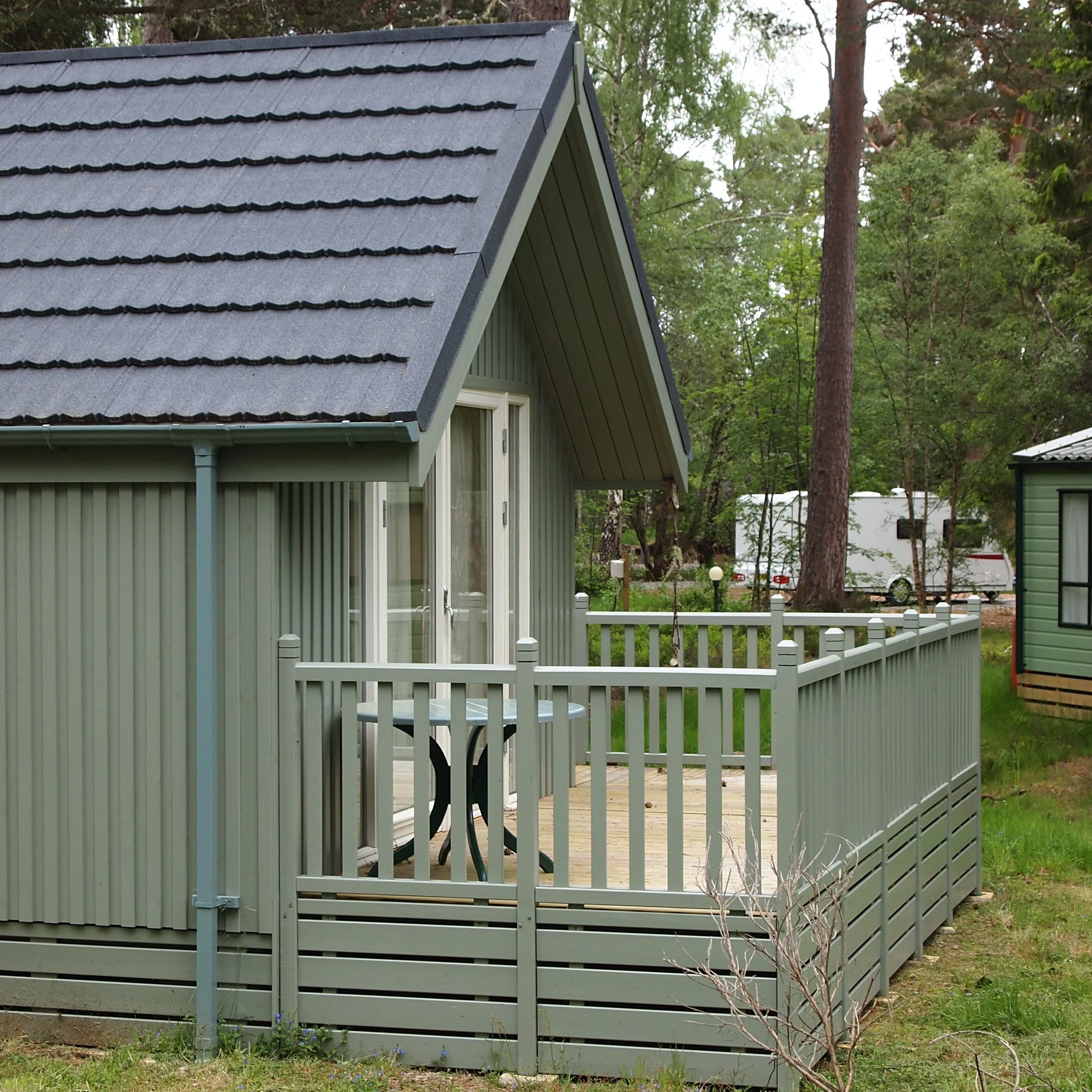 A green house with a steep gray roof and a small deck with a matching green railing, a black table, and a backyard with trees, grass, a trailer, and another house.