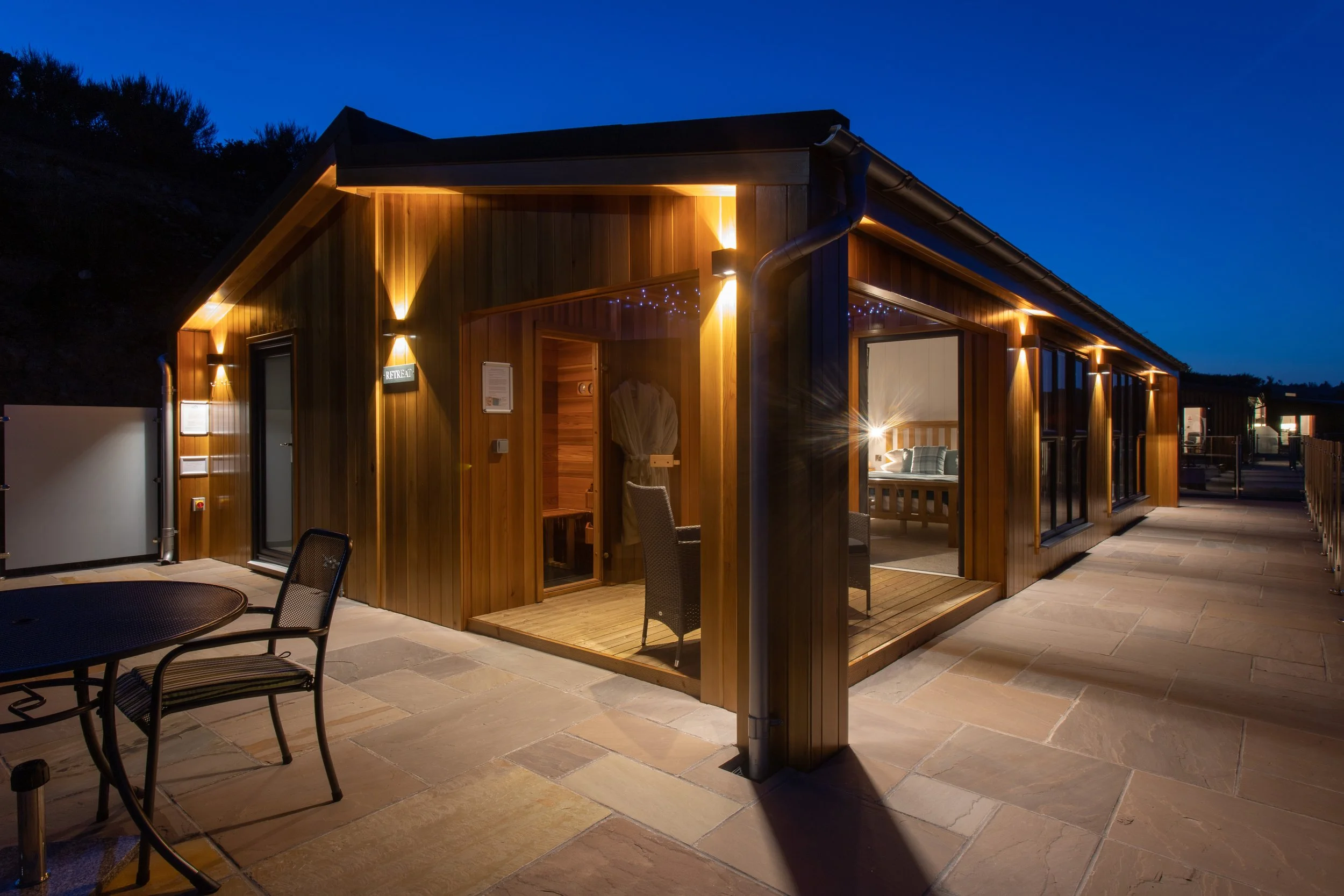 A wooden cabin illuminated by exterior lights during the evening, with a patio area including a black metal table and chairs, and large glass doors revealing a cozy bedroom inside.