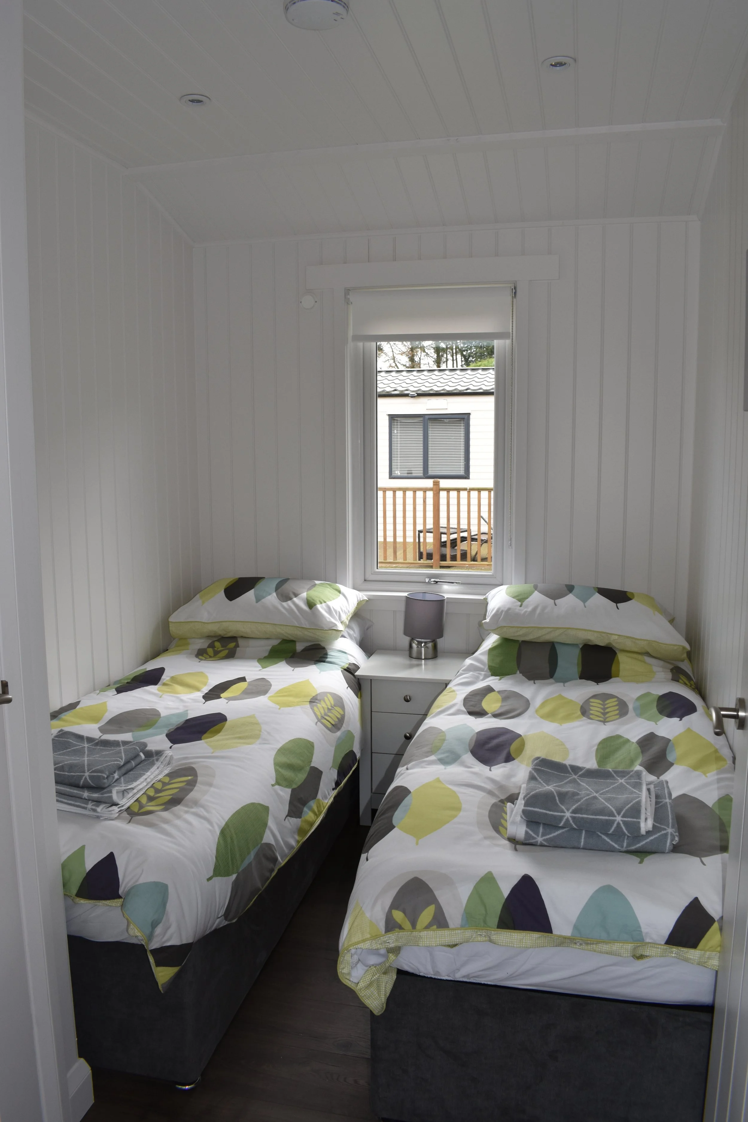A small bedroom with white paneled walls and ceiling, featuring two twin beds with colorful leaf-patterned bedding, a white nightstand with a small lamp, and a window with a view of a neighboring house and trees outside.