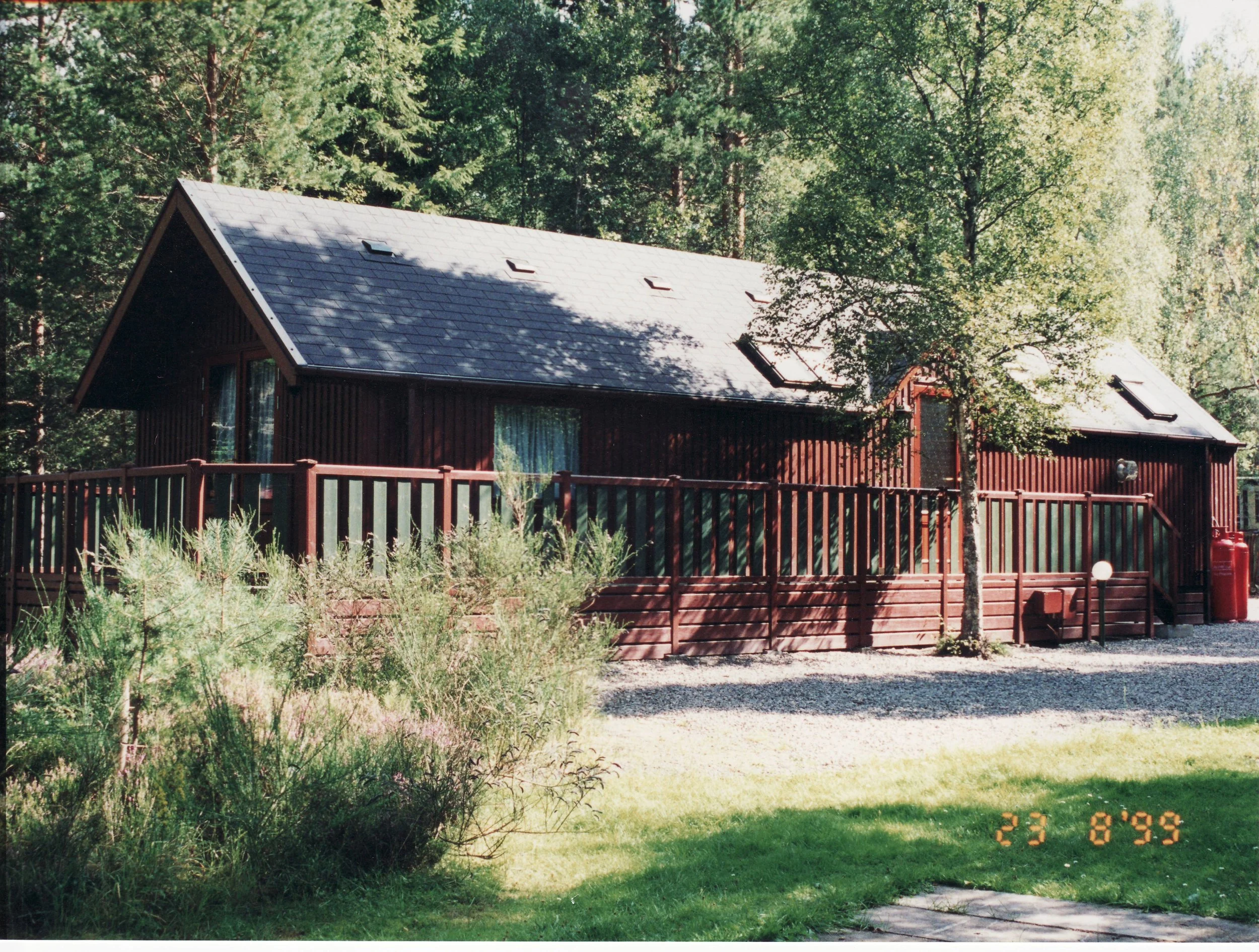 A two-story brown wooden house with a gray roof, trees surrounding it, and a yard with grass and bushes.