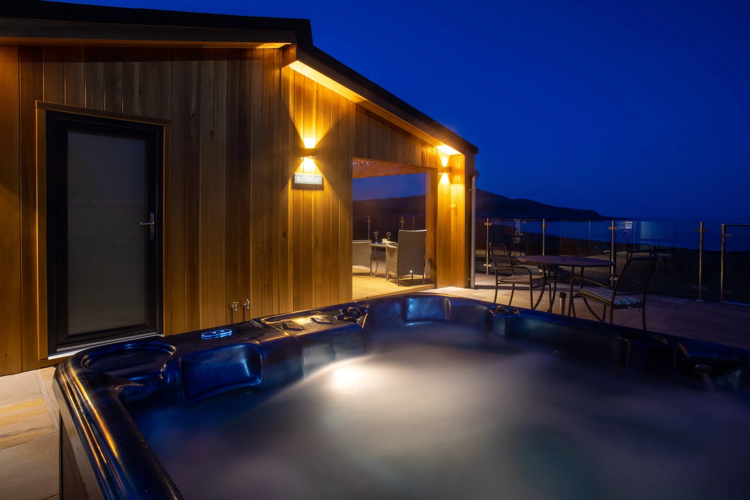 Night view of a hot tub on an outdoor balcony with a wooden house, glass railing, and mountains in the background, illuminated by warm lights.
