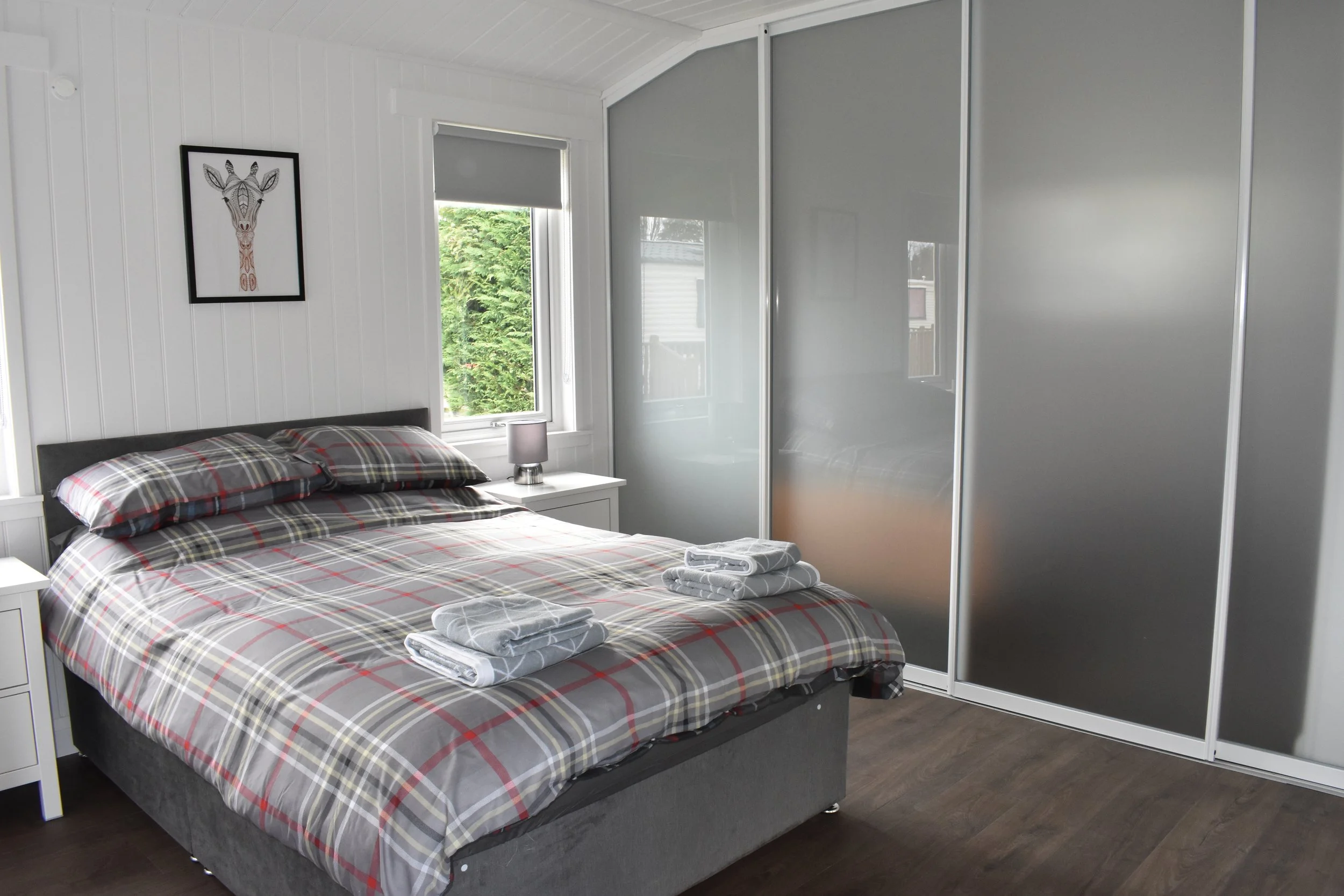 Bedroom with a bed, nightstand with lamp, window with greenery outside, closet with sliding frosted glass doors, and wall art of a giraffe