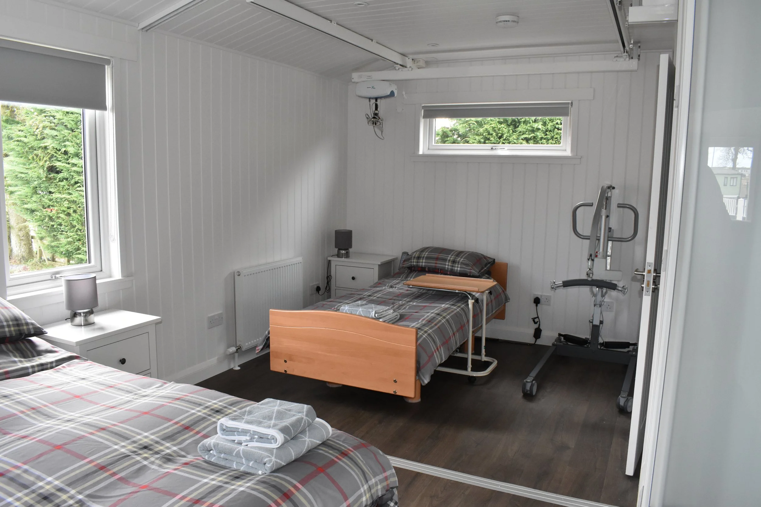 A hospital or care facility bedroom with two beds, plaid bedding, bedside tables with lamps, a window with green trees outside, a small window, a radiator, and medical equipment including a walker and bed rail