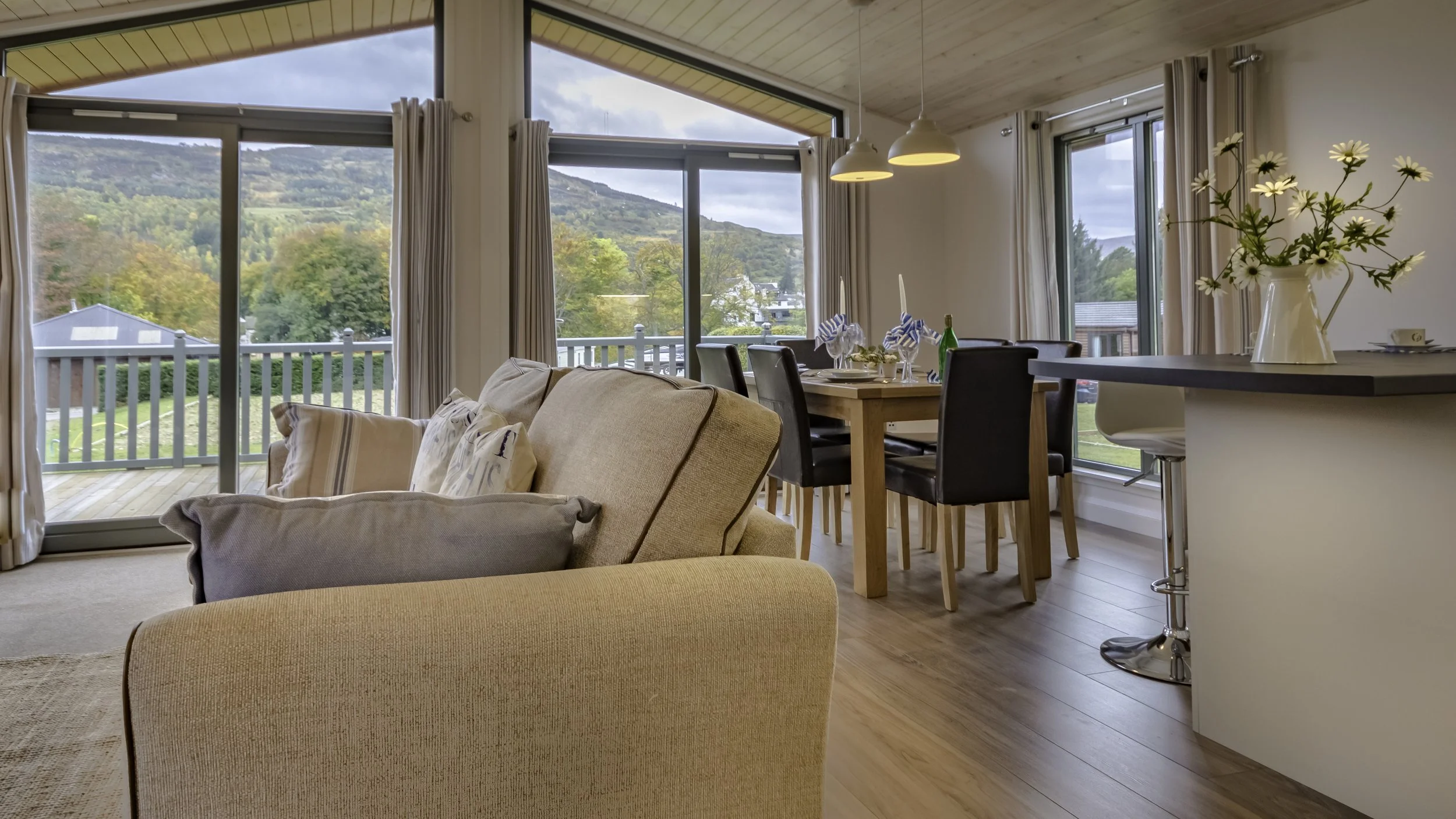 Living room with beige sofa, cushions, glass sliding doors, dining table with chairs, large windows showing a mountainous landscape, and decorative flowers in a white vase.