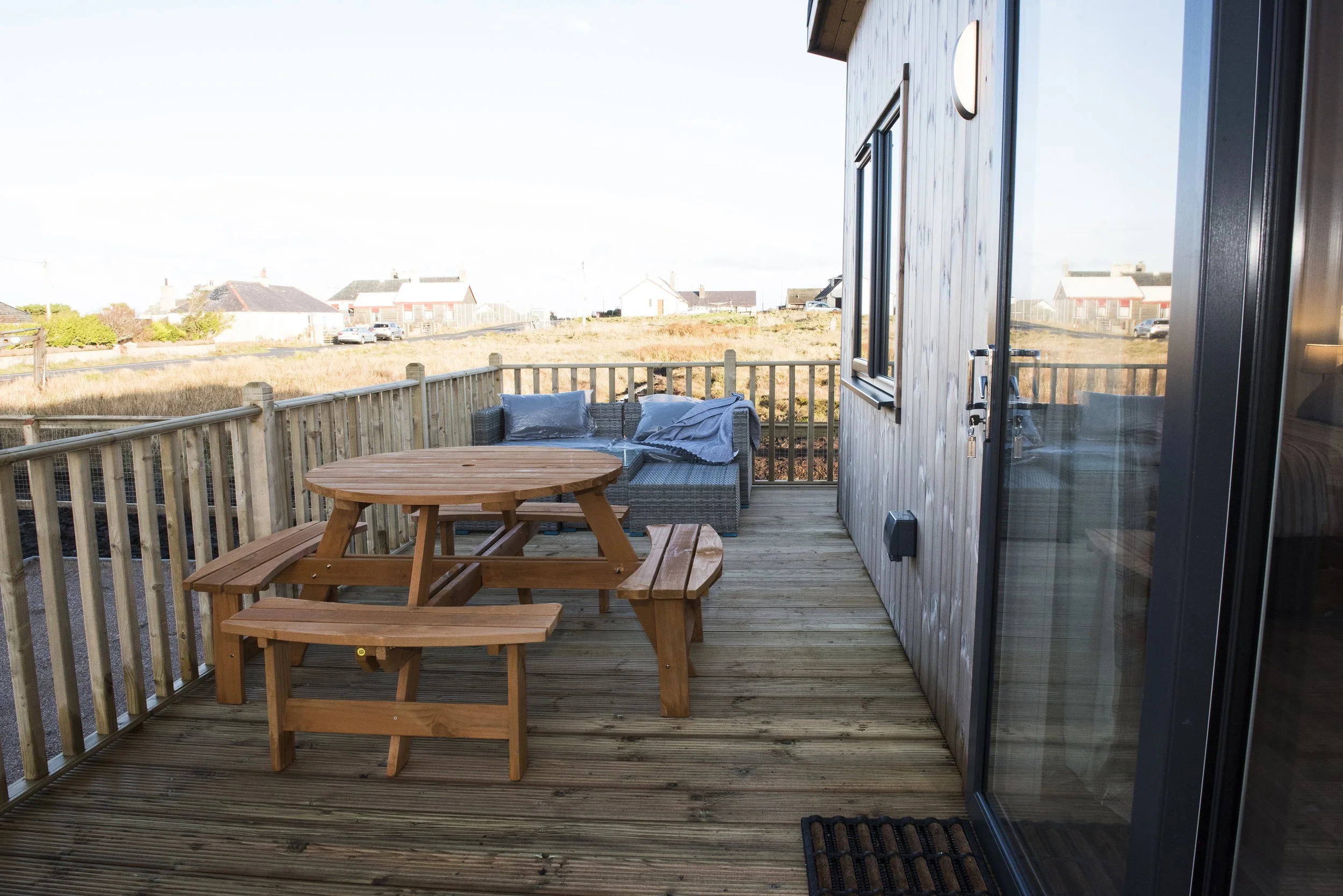 Wooden outdoor balcony with round picnic table and bench, wicker lounge corner with cushions, overlooking a grassy field with houses in the distance.