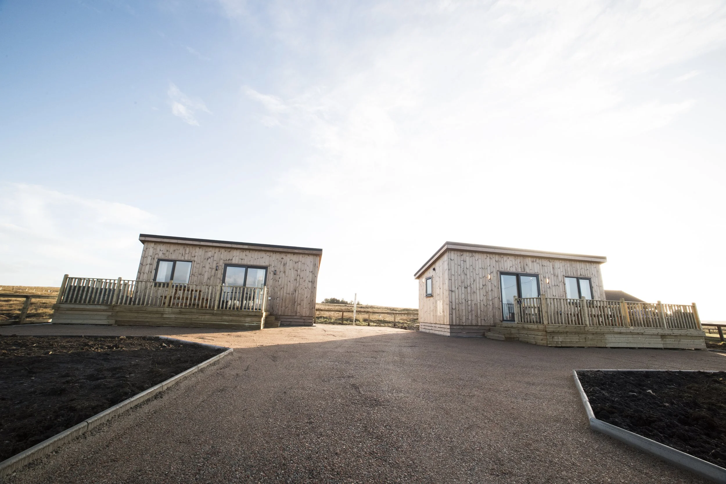 Two small wooden houses with decks face each other on a gravel driveway under a bright sky.