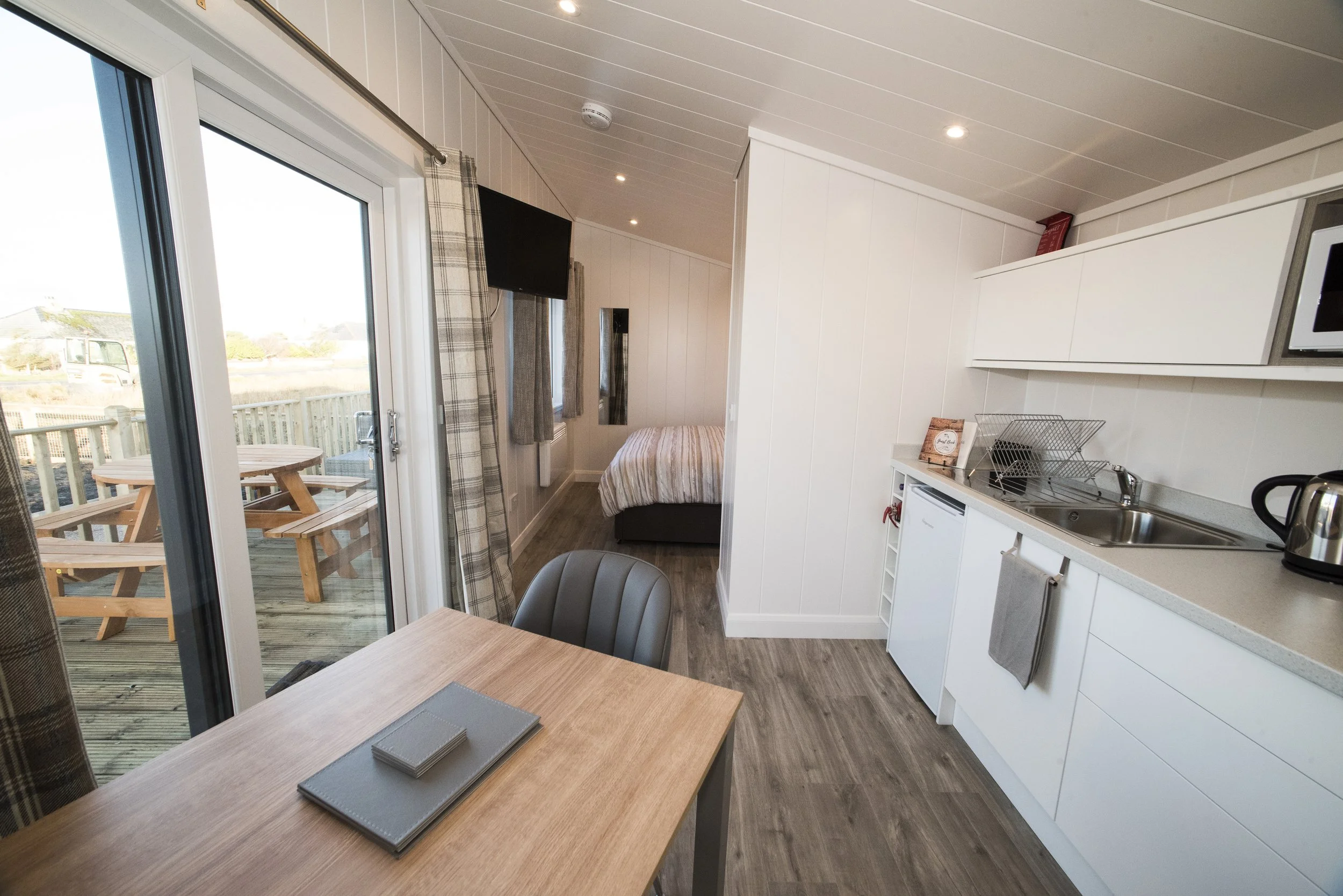 Interior of a small apartment with a kitchen, dining area, and a bedroom visible in the background. The kitchen has white cabinets, a sink, microwave, and kettle. The dining table has a gray placemat. There is a sliding glass door leading to an outdo