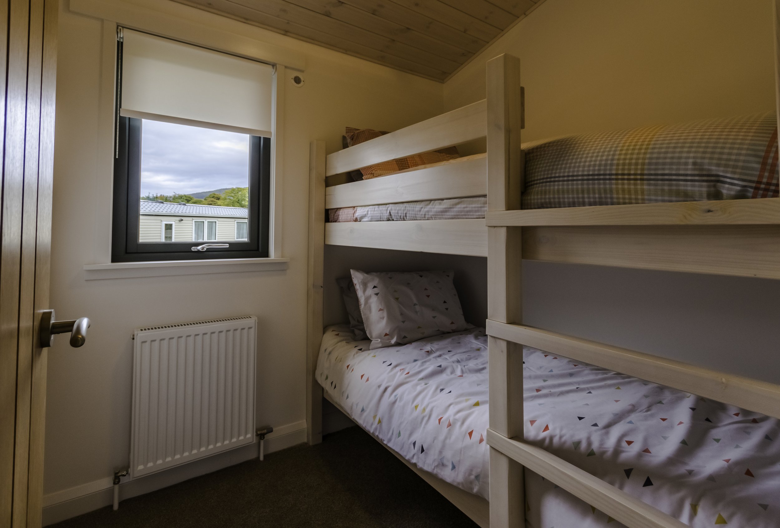 A small bedroom with a window showing a view of neighboring houses and distant hills. The room has a wooden sloped ceiling, a white radiator below the window, and a wooden bunk bed with two levels, each made with three pillows and bedding. The lower 
