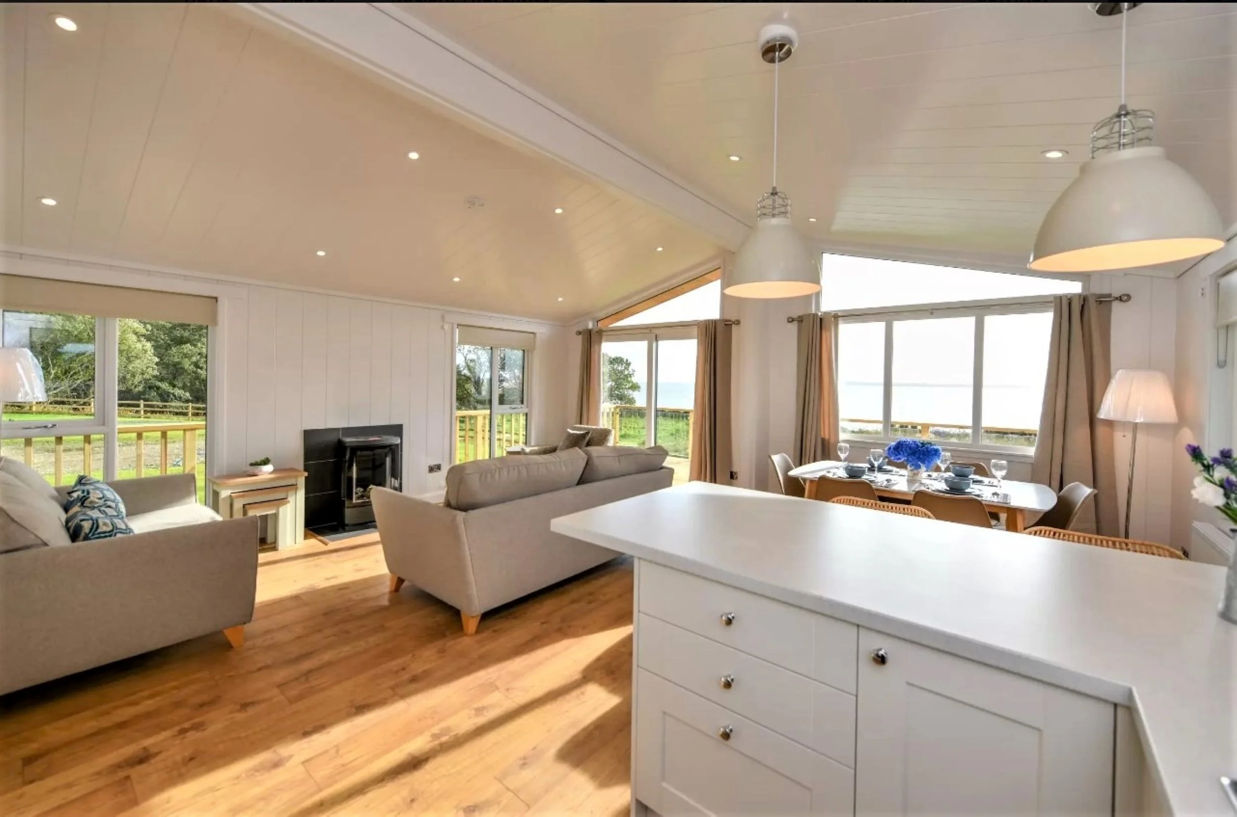 A spacious living room and dining area with large windows, beige and white furniture, hardwood floors, and modern pendant lighting.