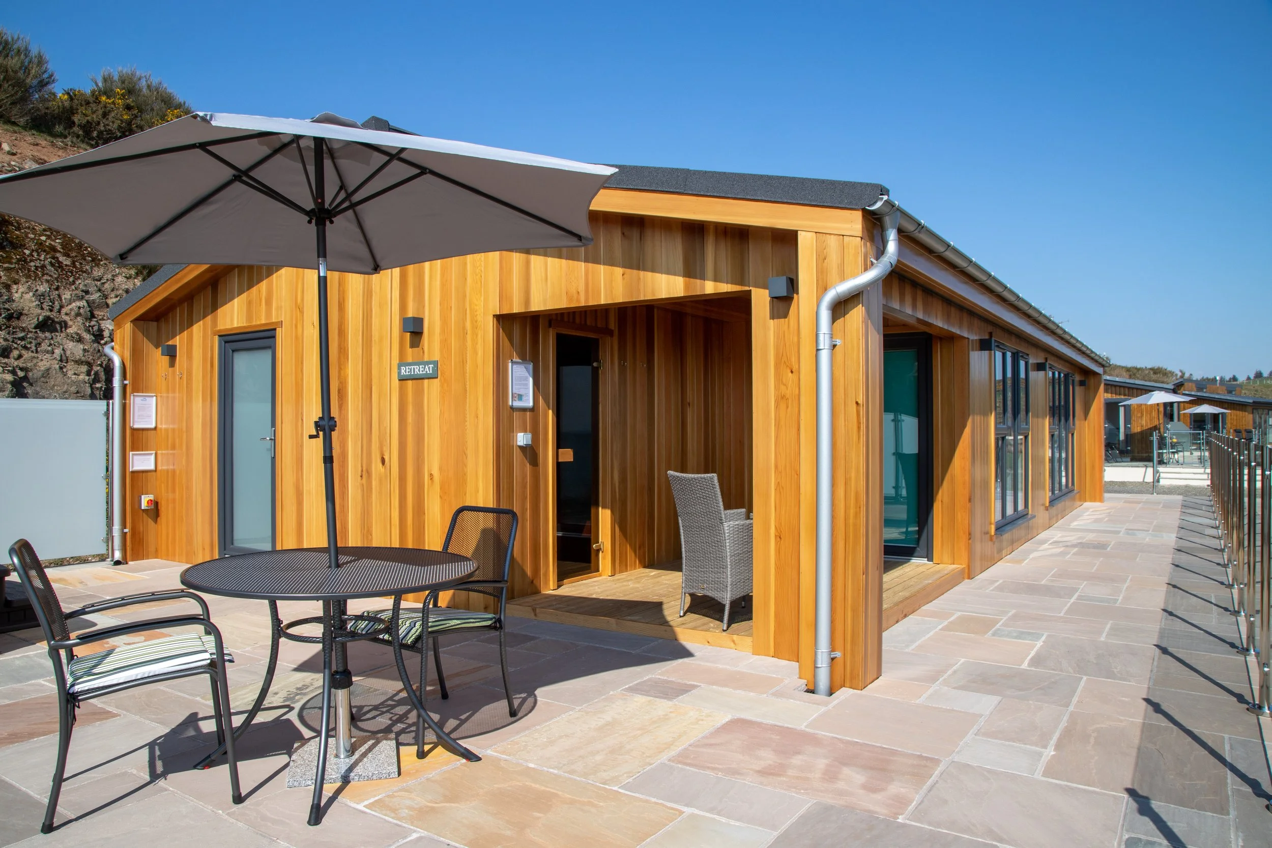 A modern wooden building with large windows and an outdoor patio featuring a round table, chairs, and a large umbrella under a clear blue sky.