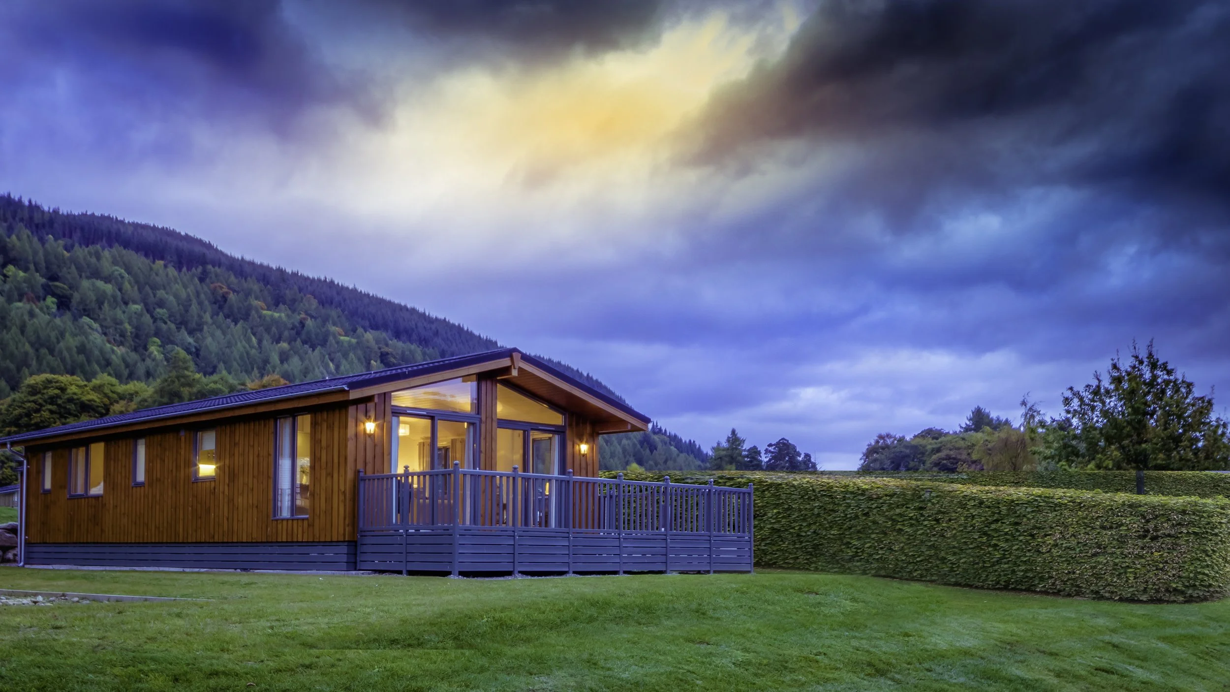 A modern wooden house with large windows and a porch, surrounded by a green lawn and trees, with a mountain and cloudy sky in the background.
