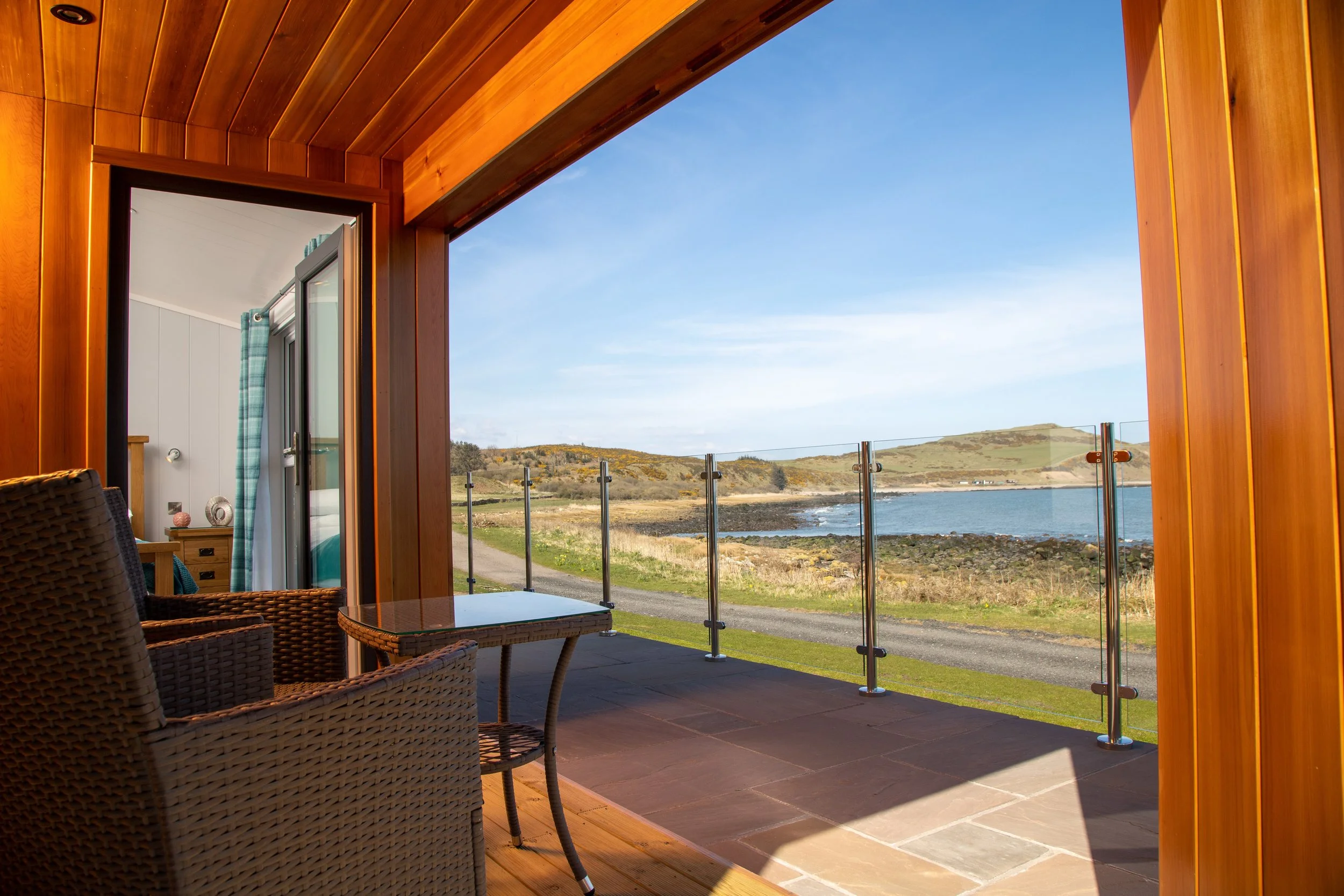 Balcony with wicker chairs and a glass table, overlooking a lake and hills under a blue sky.