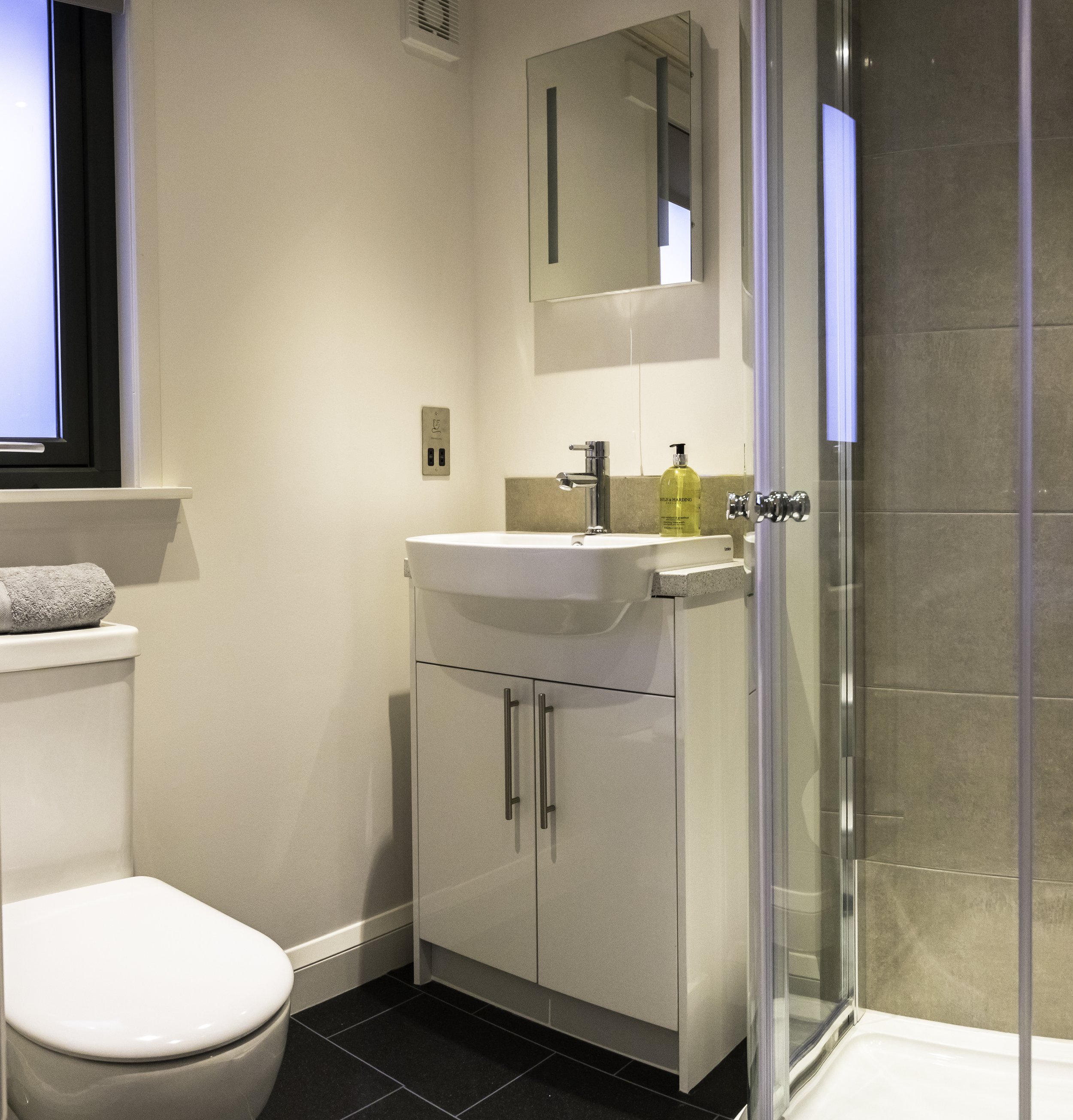 A small modern bathroom with a white toilet, a white vanity with a sink, a mirror, a yellow soap dispenser, a window, and a glass-enclosed shower with beige tiles.