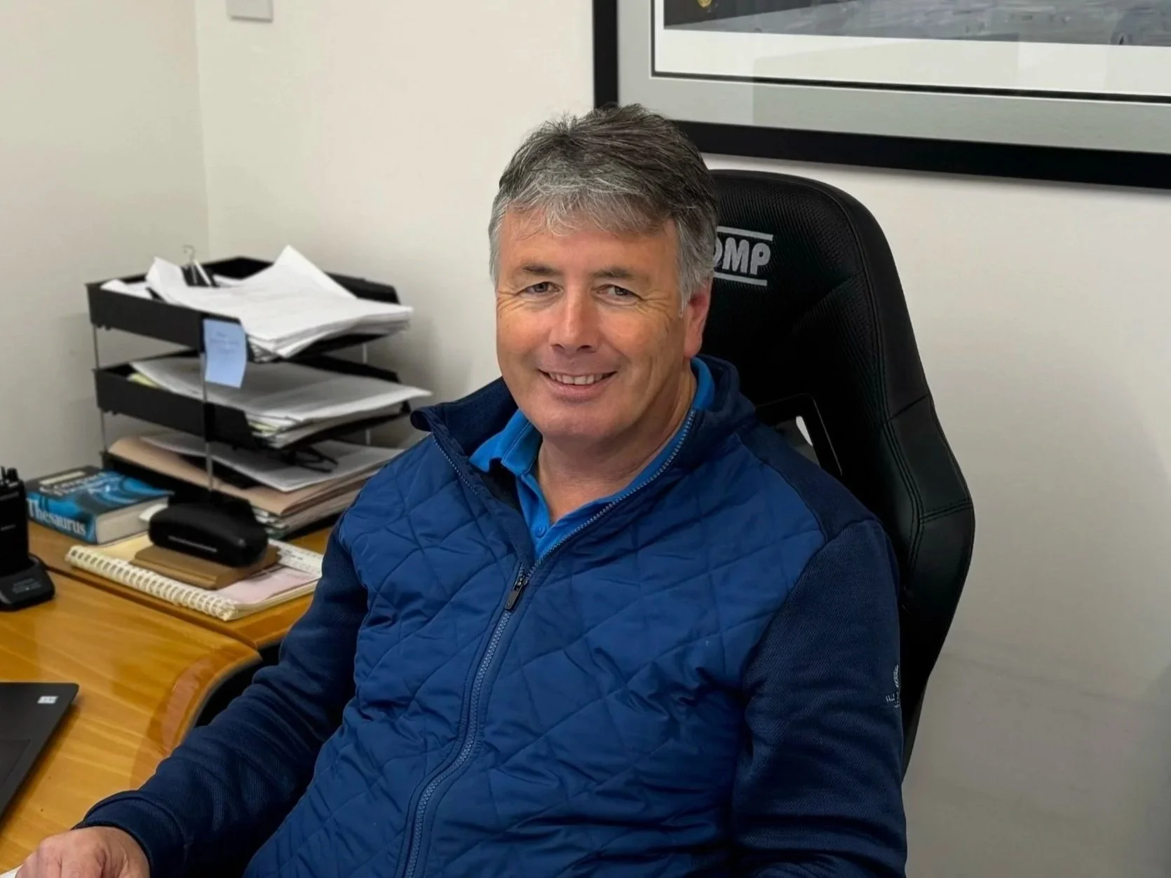 A middle-aged man with gray hair smiling while sitting in a black office chair in a cluttered office. Behind him is a desk with stacks of papers, a black tray, a telephone, a book, and notebooks. There is a framed picture or poster on the wall.