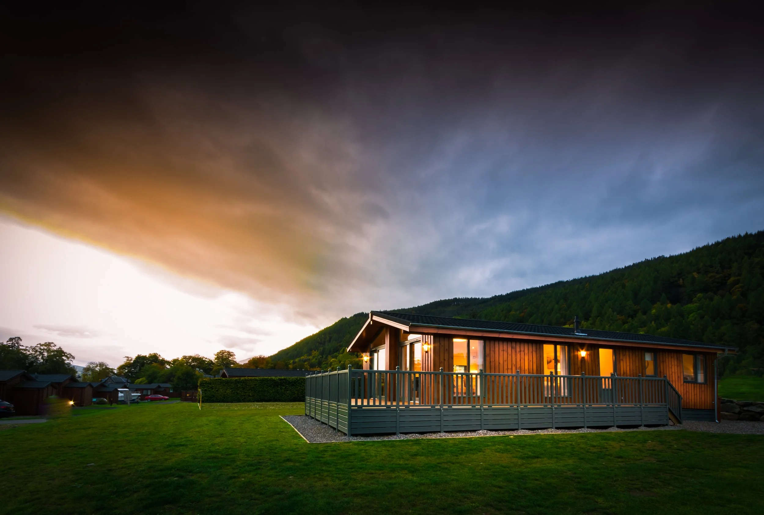 A wooden house with large windows and a deck, illuminated from inside, situated on a grassy lawn surrounded by trees and hills, under a cloudy evening sky.