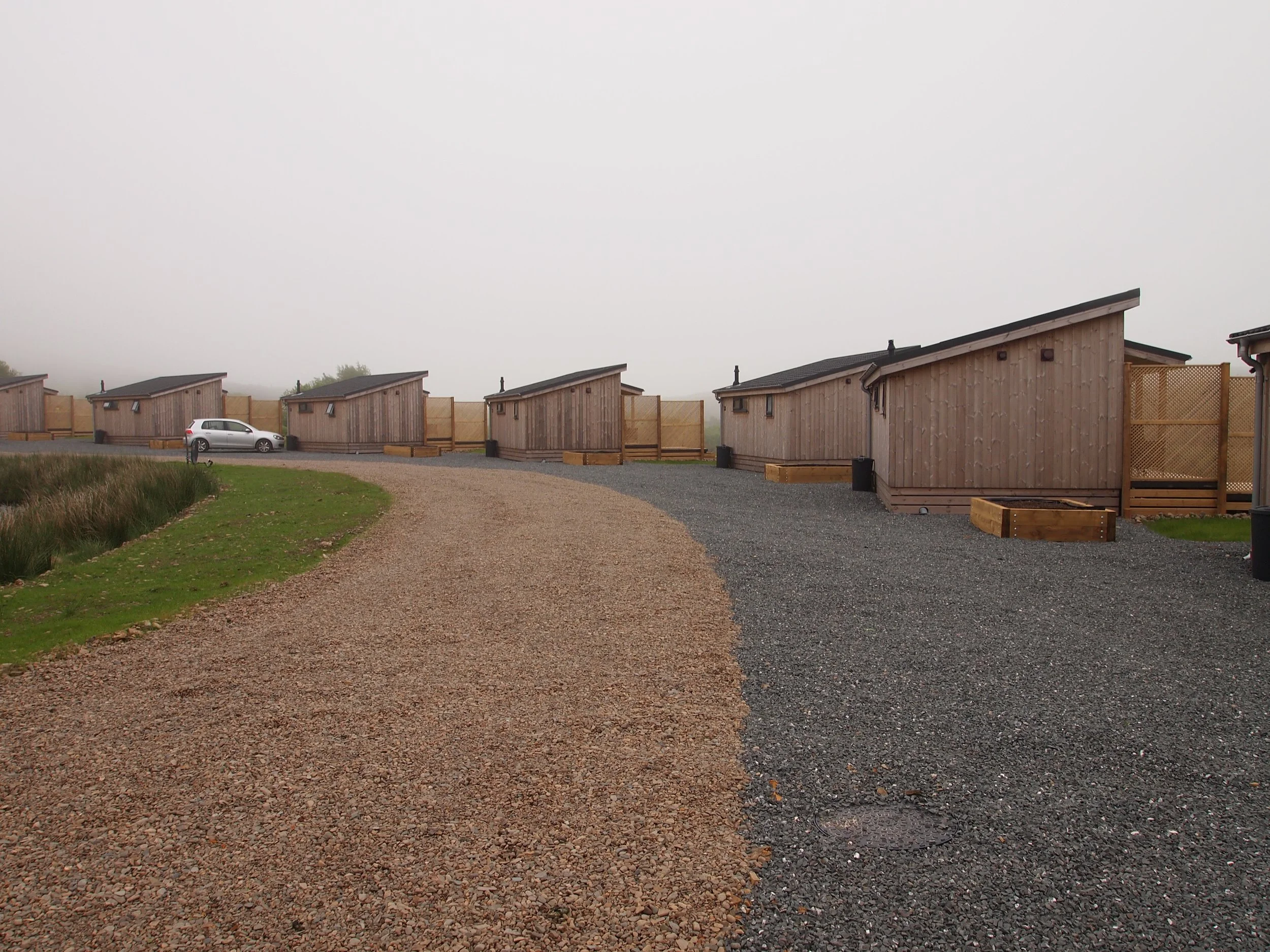 A row of small, modern wooden cabins with sloped roofs lined along a gravel pathway in a foggy outdoor setting.