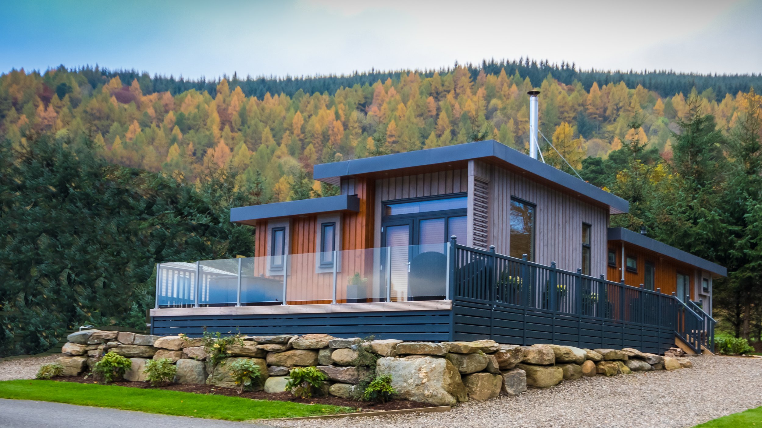 Modern house with wooden siding and a large deck, built on rocky terrain, surrounded by trees and set against a forested hillside.