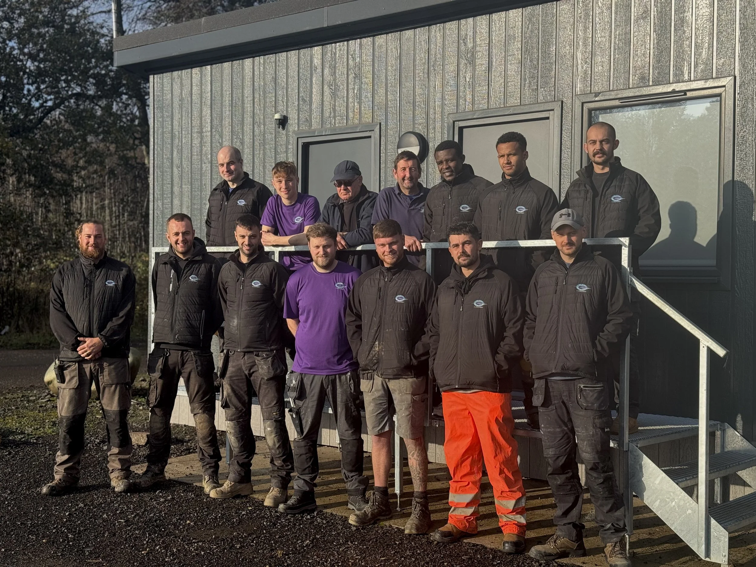 Group of 15 men standing outside in front of a gray building, some on stairs and others on ground, wearing work uniforms including black jackets, purple shirts, and high-visibility pants, with trees and sky in the background.