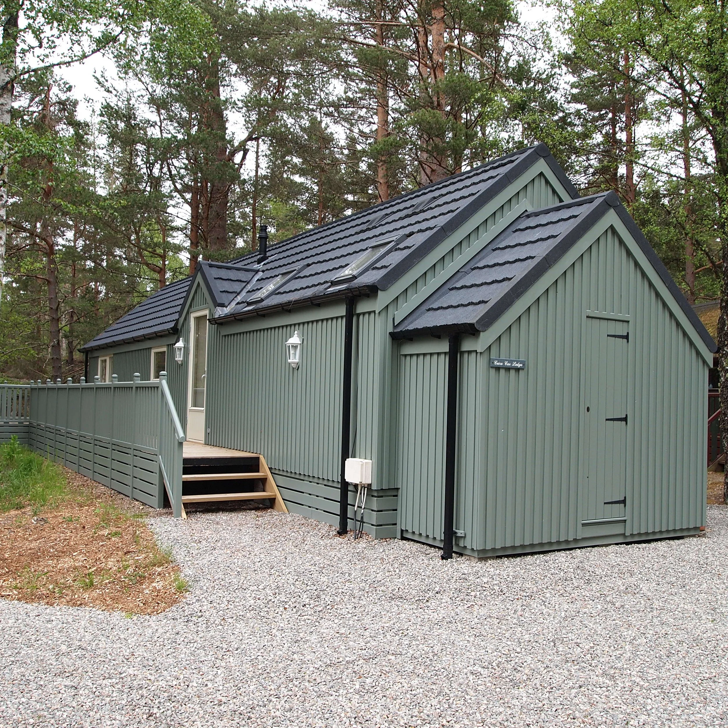 A green wooden house with black roof shingles and skylights, surrounded by trees, with a gravel driveway in the foreground.