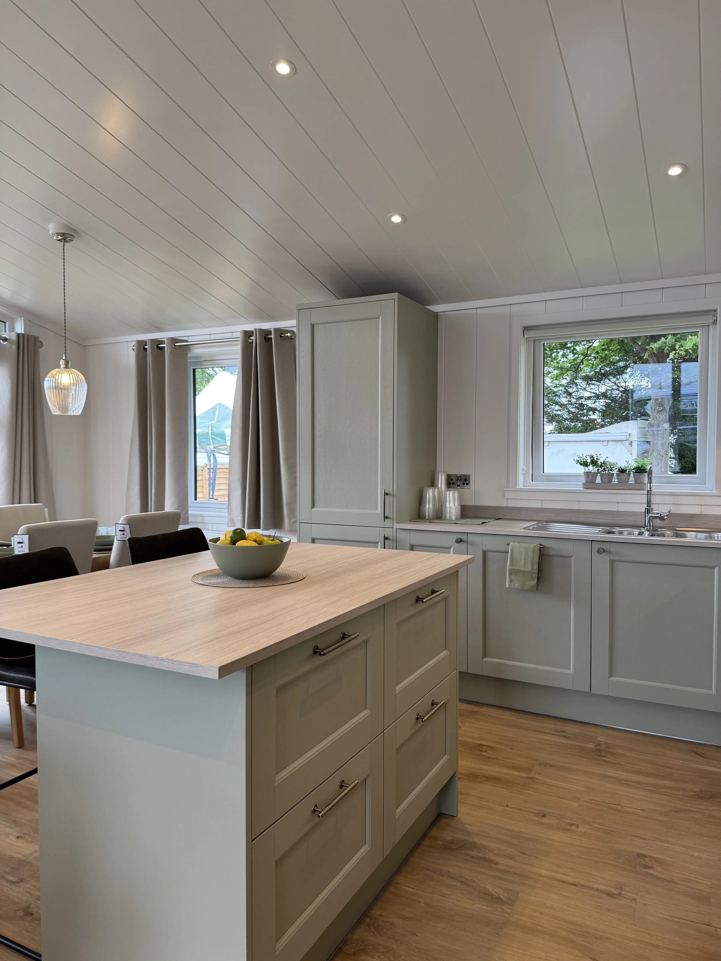 A modern kitchen with light gray cabinets, a wooden kitchen island with drawers, a bowl of lemons on the island, windows with beige curtains, and a hardwood floor.