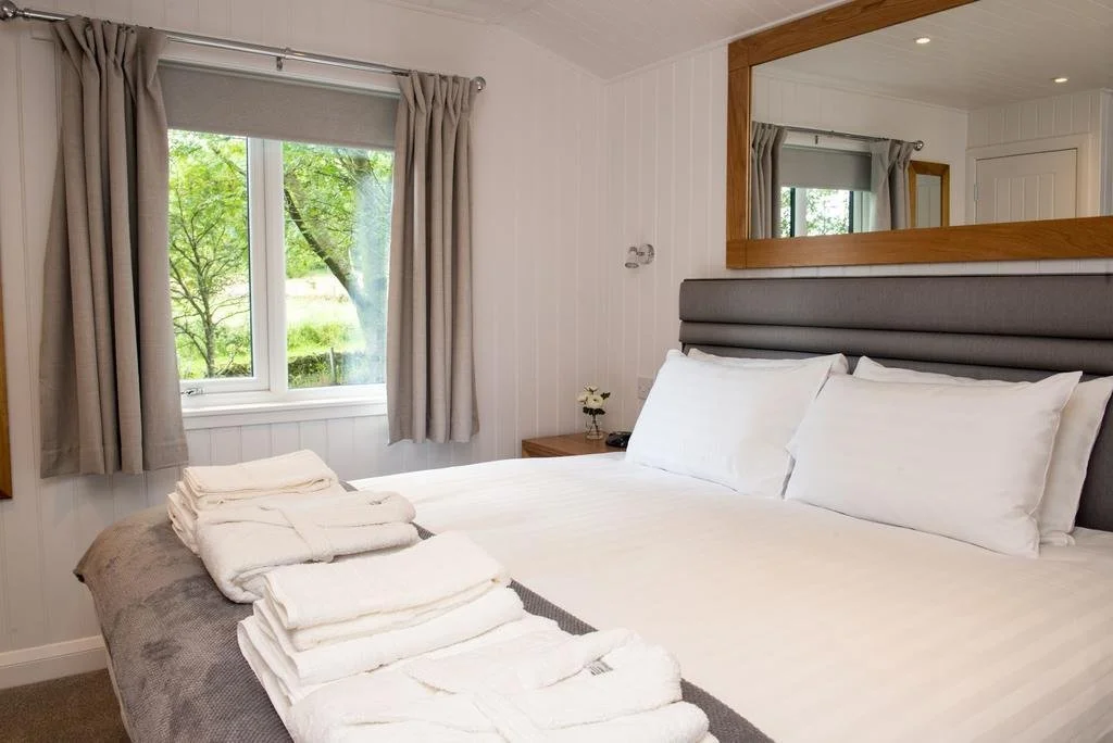 A neatly made bed with white linens and multiple pillows in a cozy, well-lit room with a large window and cream curtains, overlooking greenery outside.