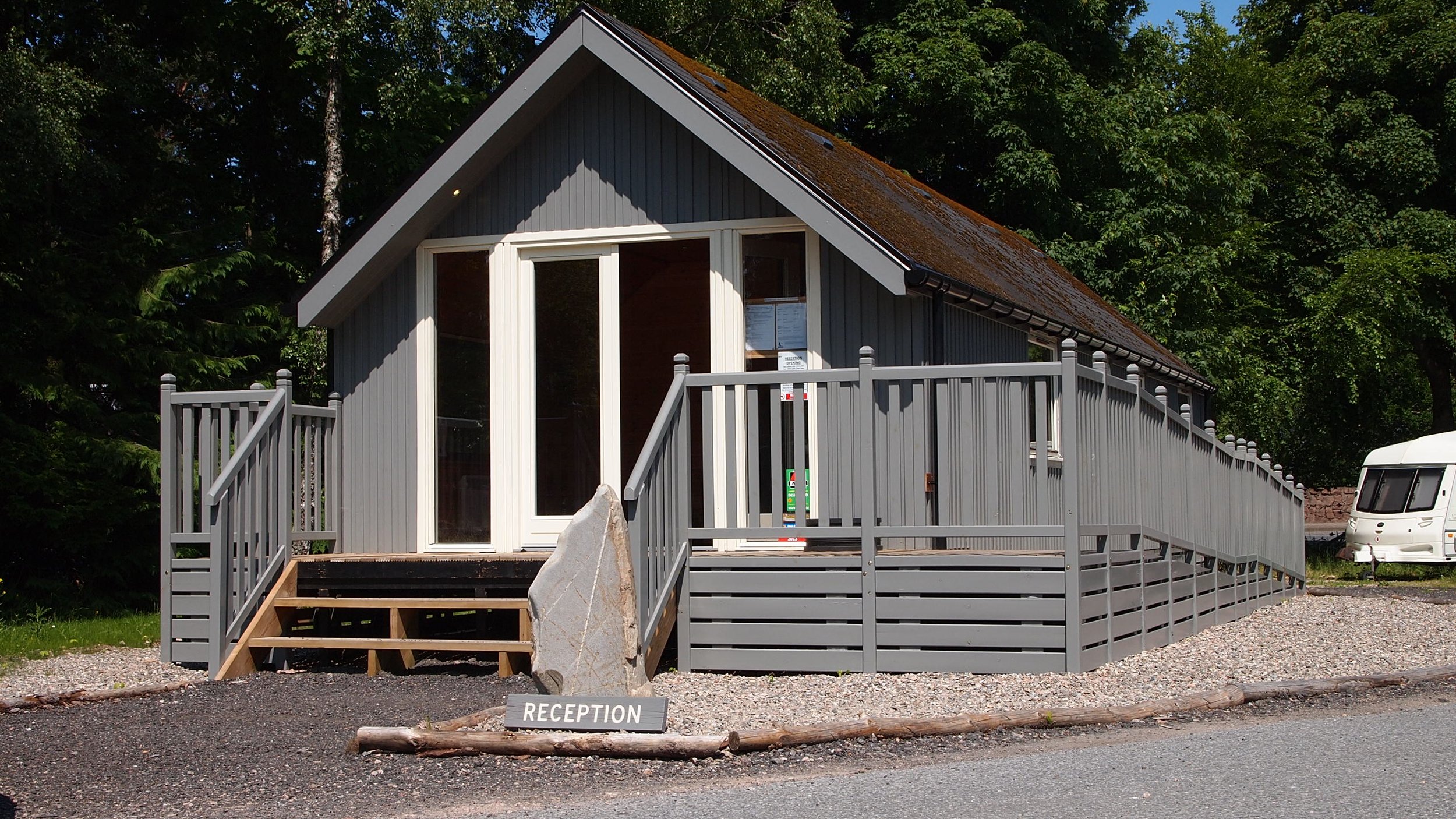 Gray wooden reception building with a ramp, staircase, large glass doors, and surrounded by green trees. There is a sign that reads 'RECEPTION' in front of the building and a white caravan is parked to the right.
