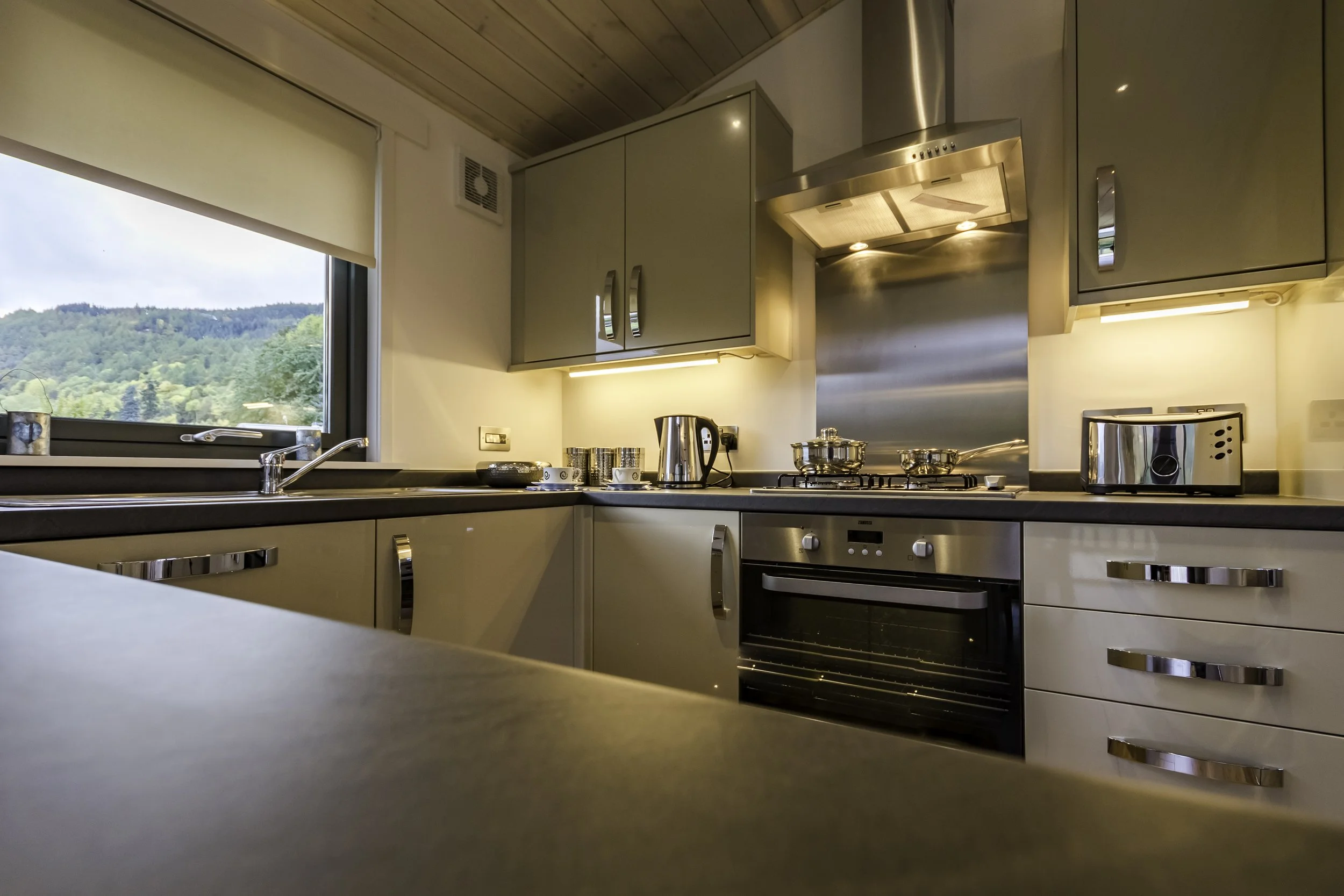 Modern kitchen with beige cabinets, stainless steel appliances, black countertop, window with outdoor view, and wooden ceiling.