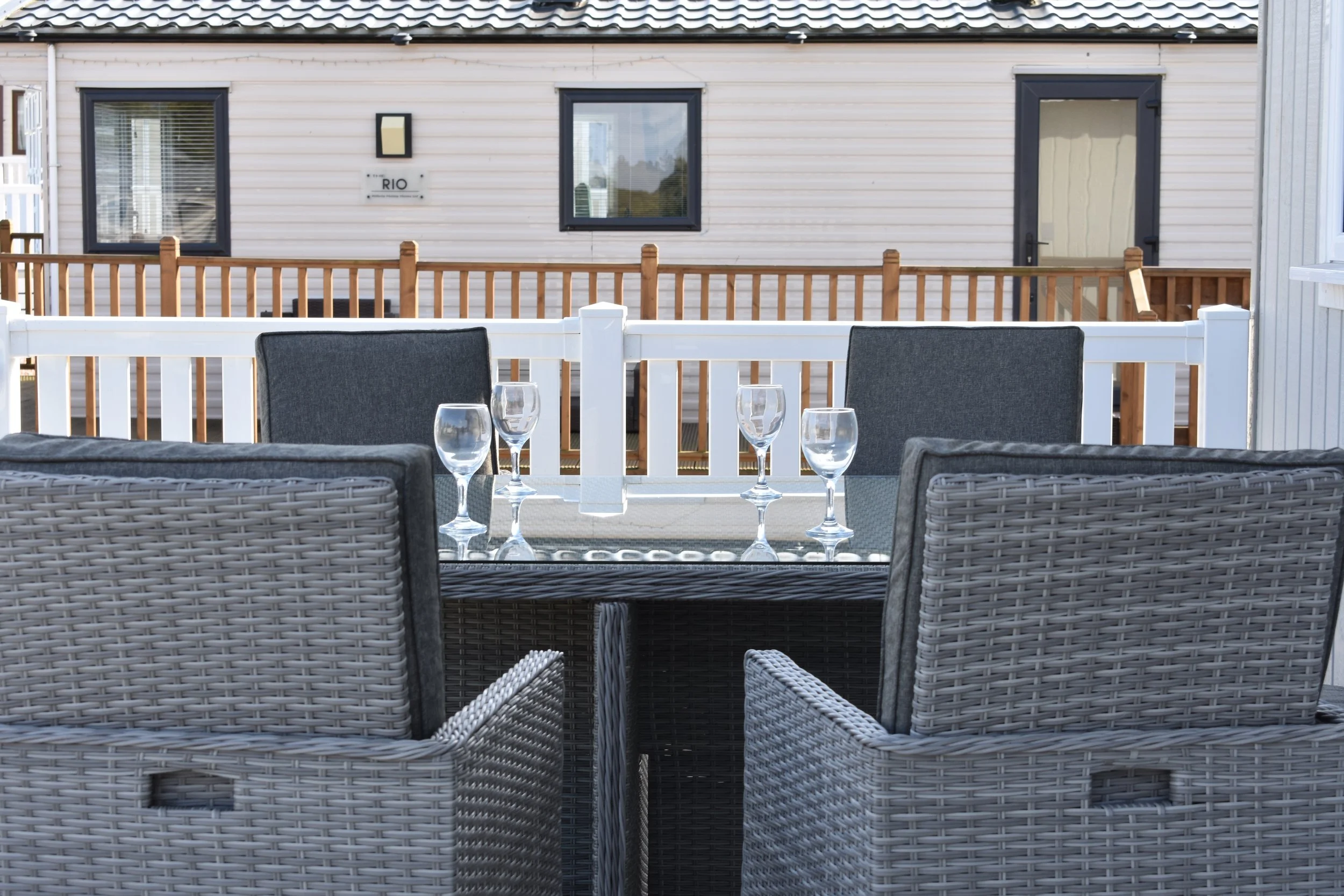 Outdoor patio table set with four wine glasses, surrounded by wicker chairs, on a deck with a wooden railing, in front of a beige house with black-trimmed windows and a door.